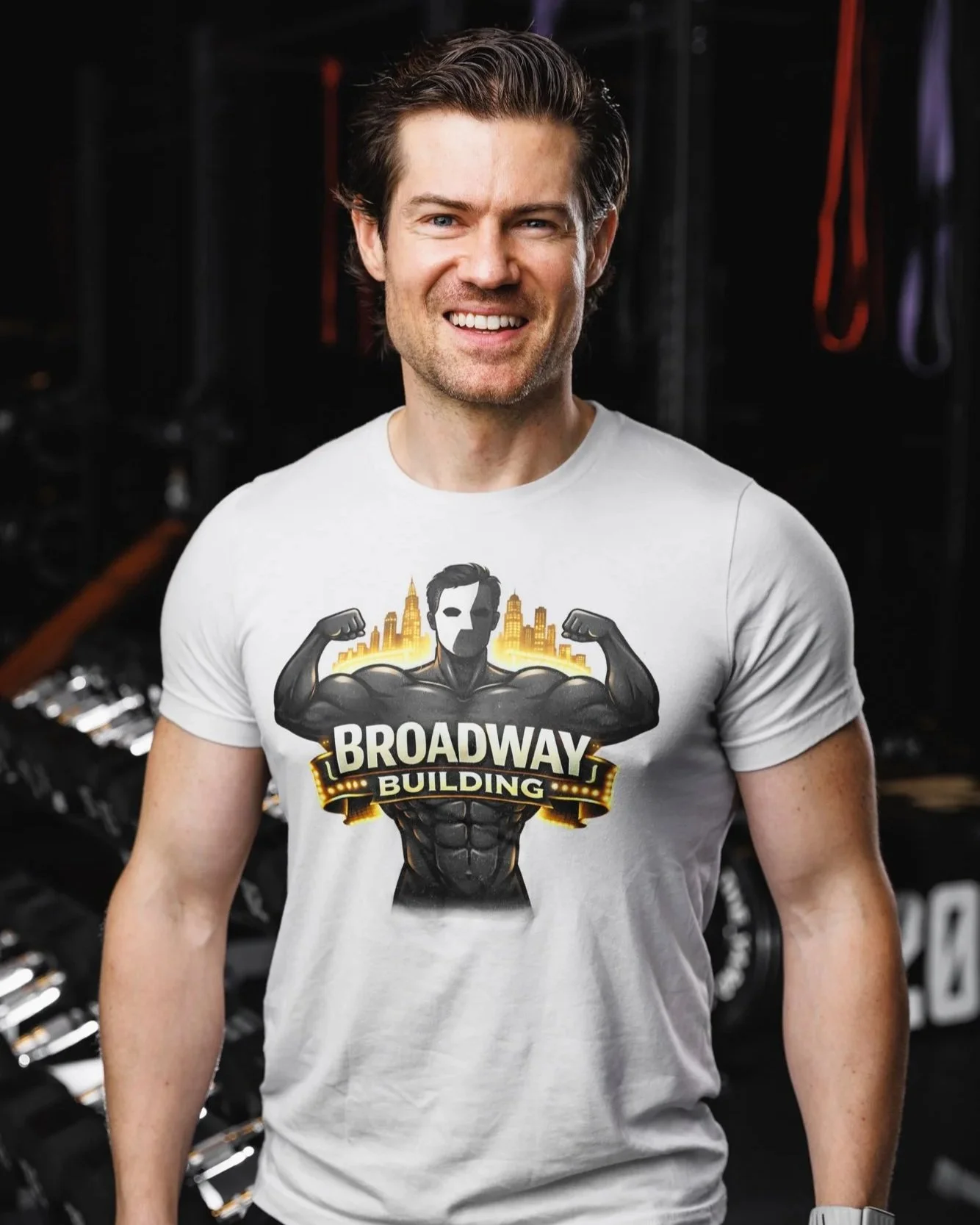 A man with short dark hair and a big smile wearing a white T-shirt with a graphic of a muscular superhero and the words 'BROADWAY BUILDING' in a gym or fitness setting.