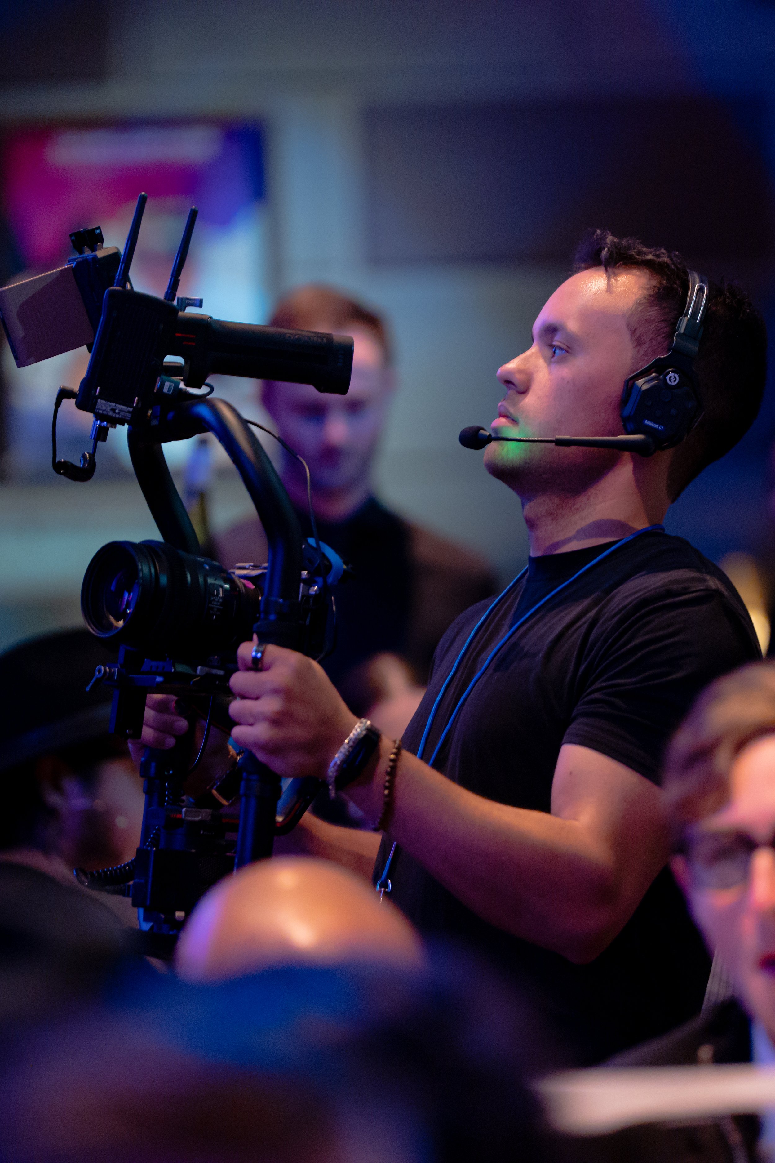 A man operating a professional video camera at an indoor event, wearing a headset with a microphone.