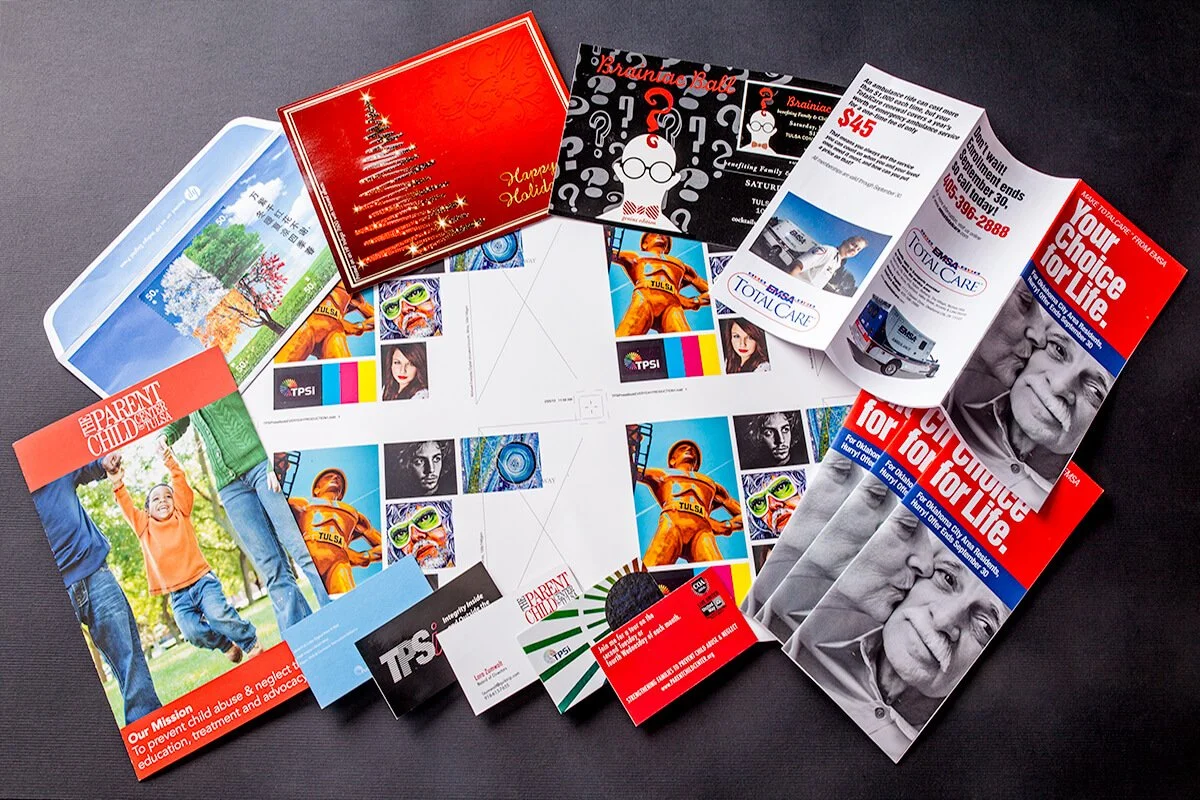 Collection of flyers, brochures, and promotional materials for community organizations and services, including Christmas-themed cards and campaign literature, spread out on a dark surface.