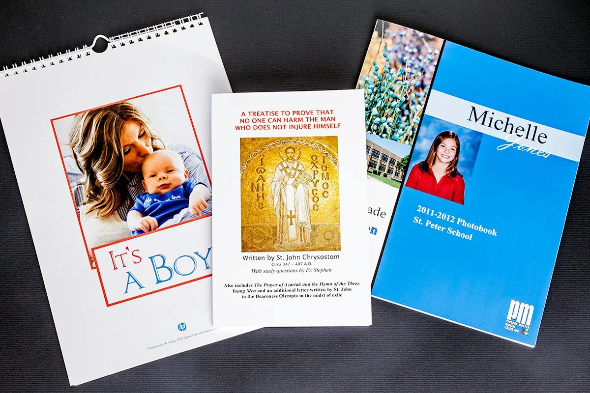 Four booklets or pamphlets are displayed on a black surface. The first on the left has a cover photo of a woman holding a baby and text that reads 'It's A Boy'. The second features religious imagery and text about a treatise by St. John Chrysostom. The third has a nature photo with a school building and a person. The fourth is a blue school yearbook cover for Michelle Jones from St. Peter School, 2011-2012.