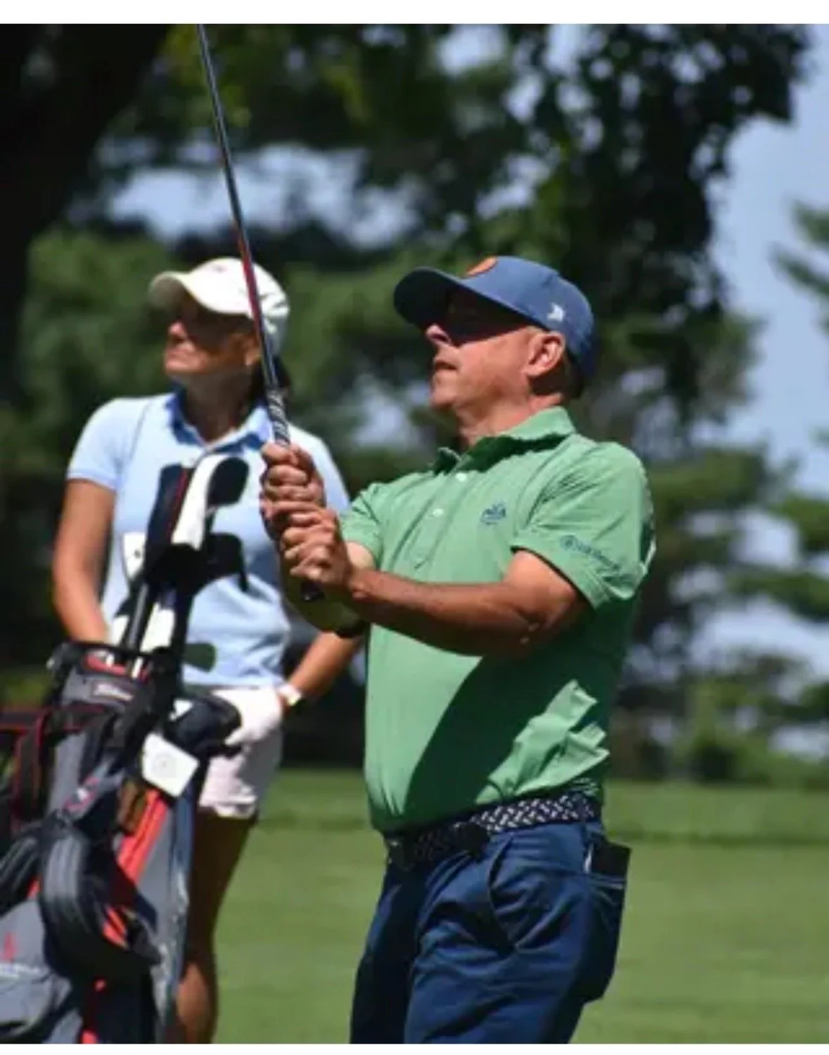 Two people on a golf course, one man wearing a green shirt and a cap, holding a golf club, and a woman watching in the background with a golf bag.