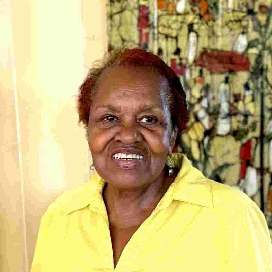 Portrait of an elderly woman smiling, wearing a yellow shirt. Background features a patterned curtain or tapestry.