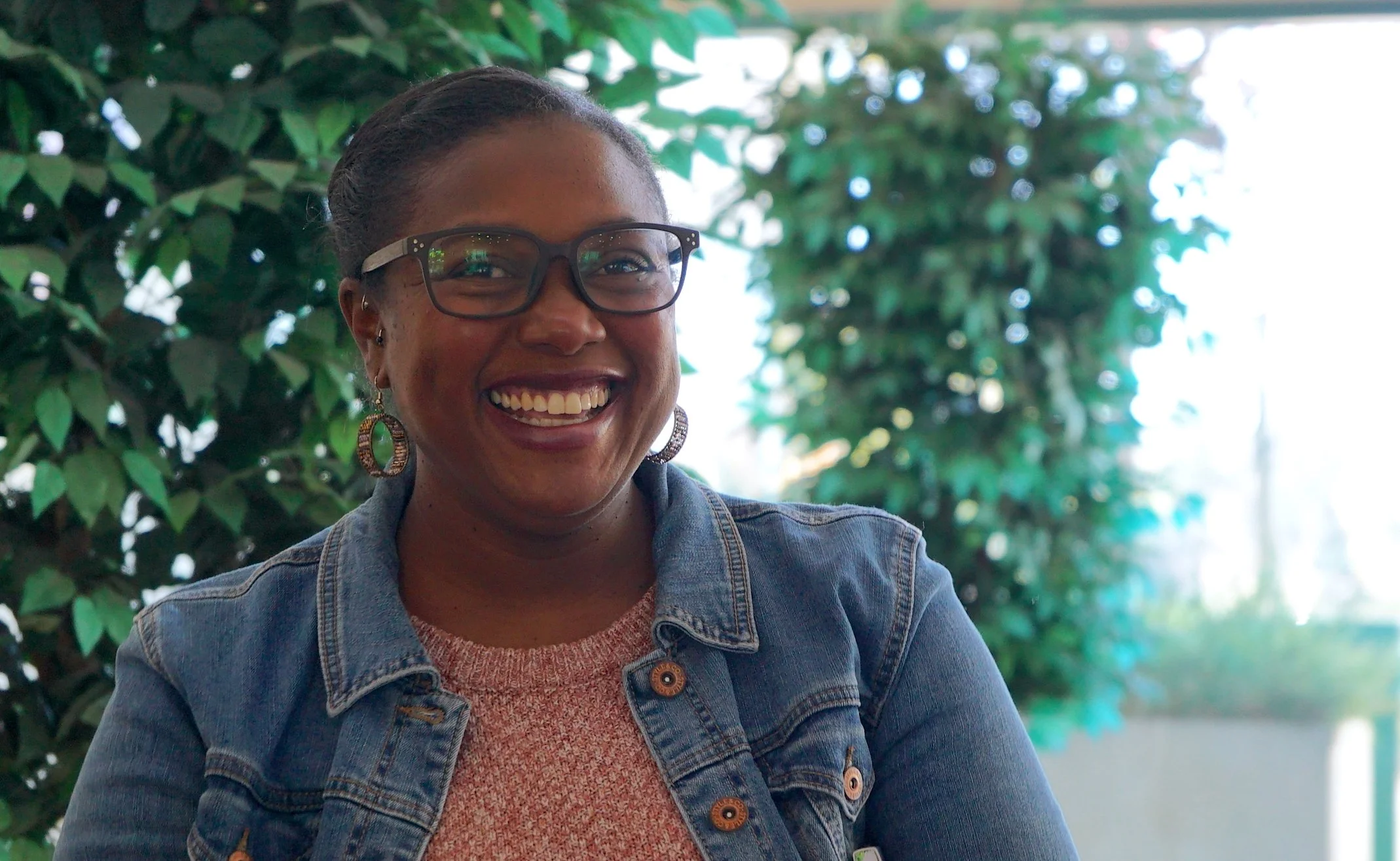 A woman with glasses smiling, wearing earrings, a pink sweater, and a denim jacket, outdoors with green foliage in the background.