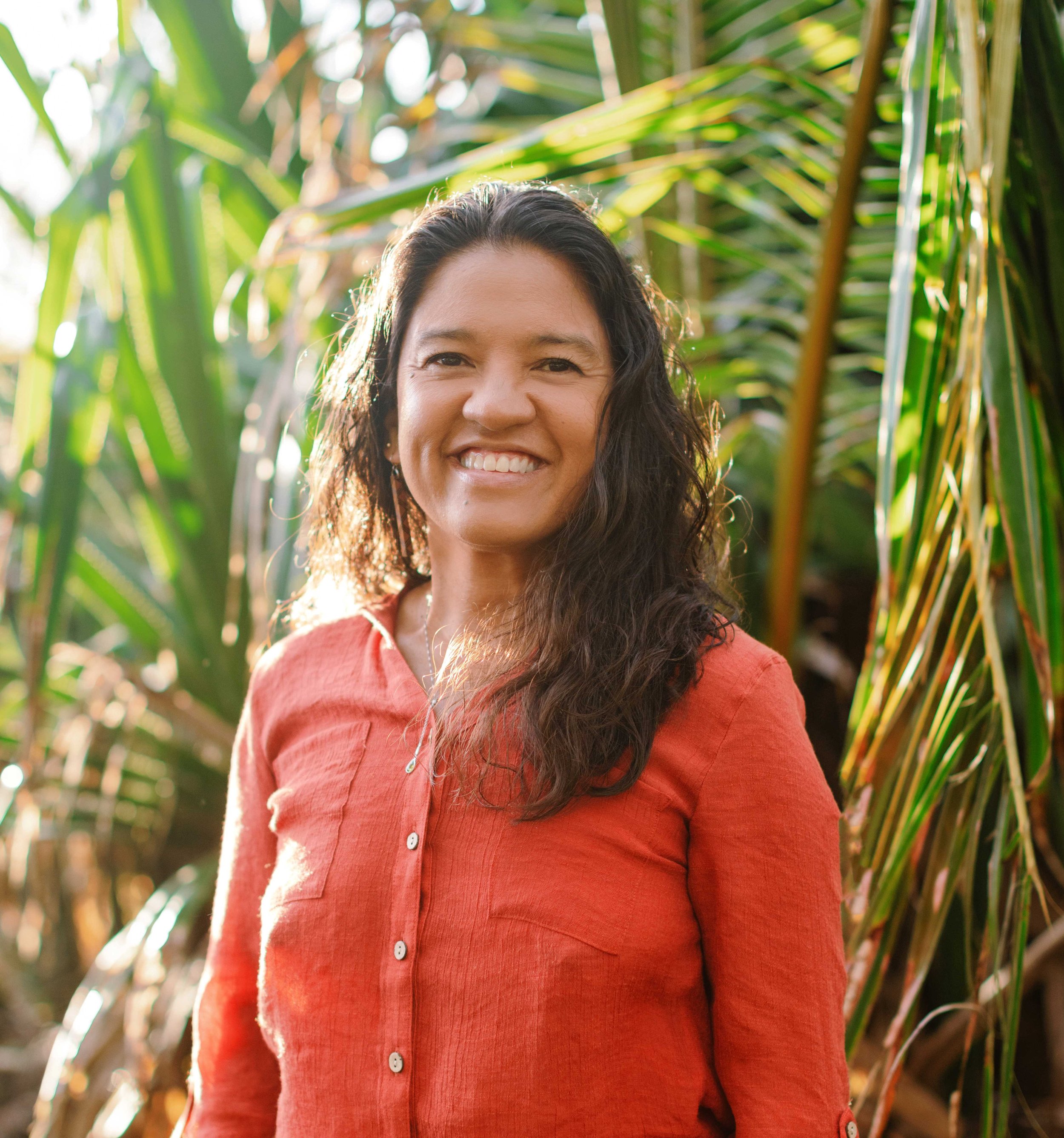 A woman with dark curly hair and wearing a coral button-up shirt, smiling outdoors in a lush green tropical setting.