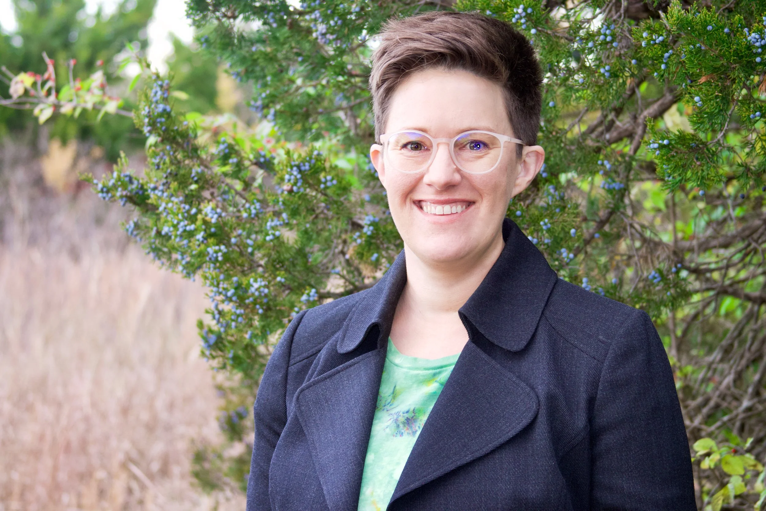 A woman with short brown hair, glasses, and a smile, standing outdoors in front of a bush with small blue flowers. She is wearing a dark coat over a green patterned shirt.