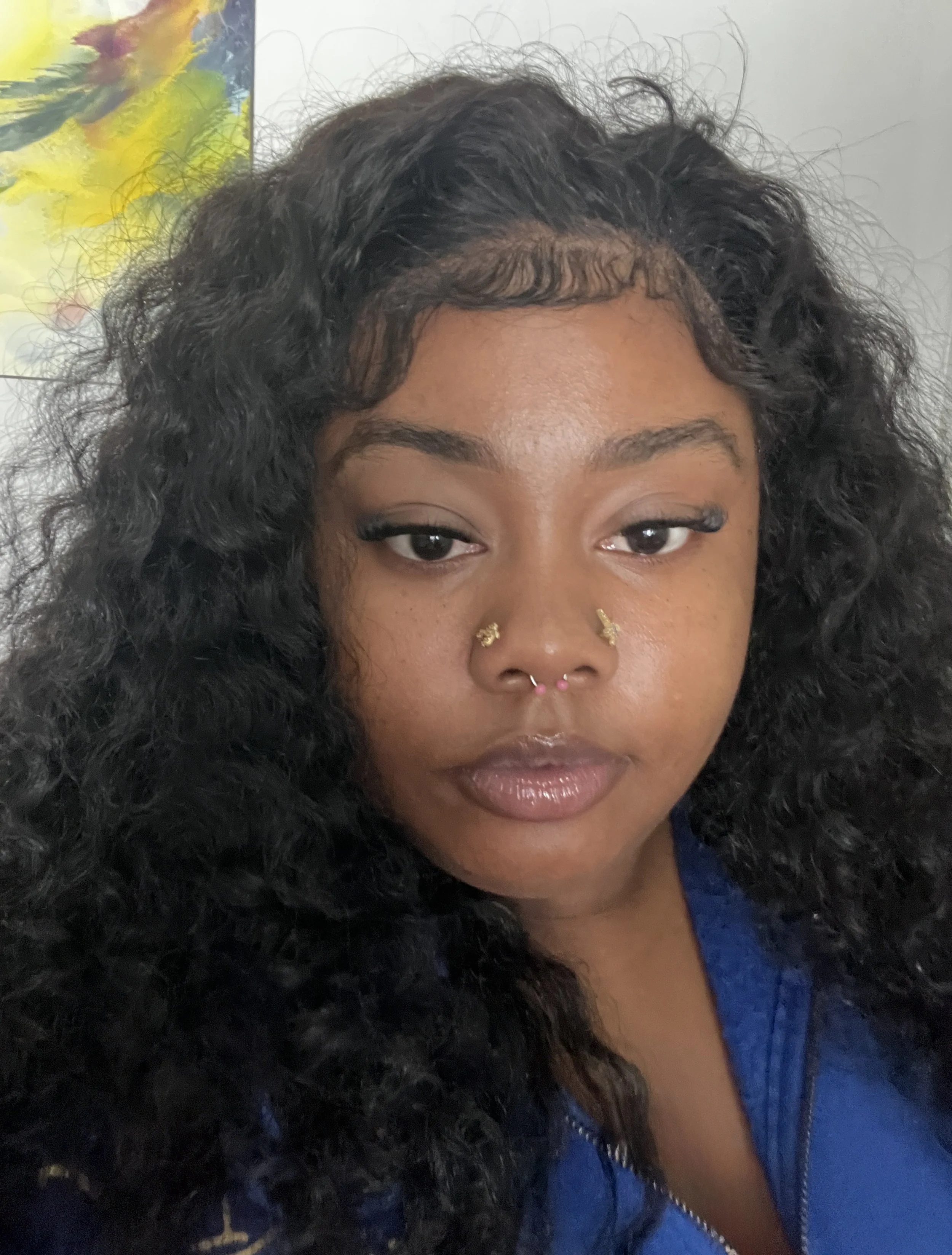 Close-up of a woman with curly black hair, wearing a blue top, with facial piercings including a nose ring and septum ring, and light makeup, standing in a room with an abstract colorful painting in the background.