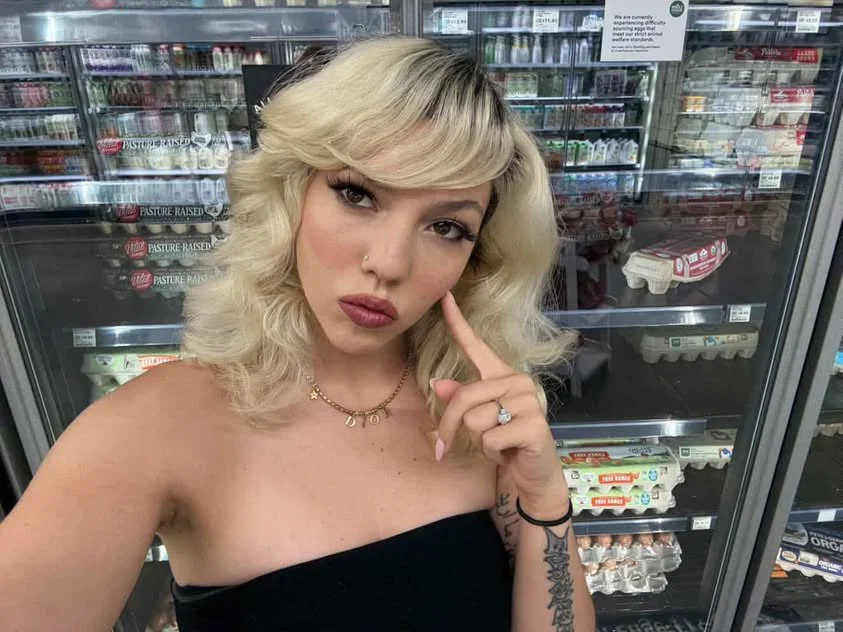 A woman with blonde wavy hair taking a selfie in a grocery store, standing in front of refrigerated shelves with dairy products.