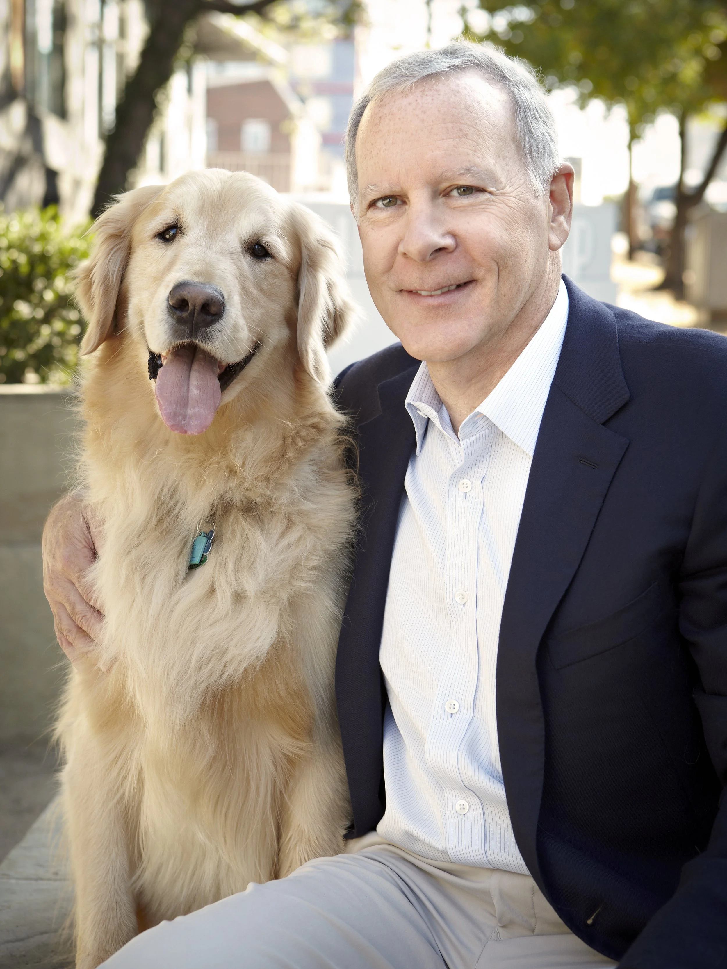 A man in a navy blazer and white shirt sitting next to a golden retriever outdoors.