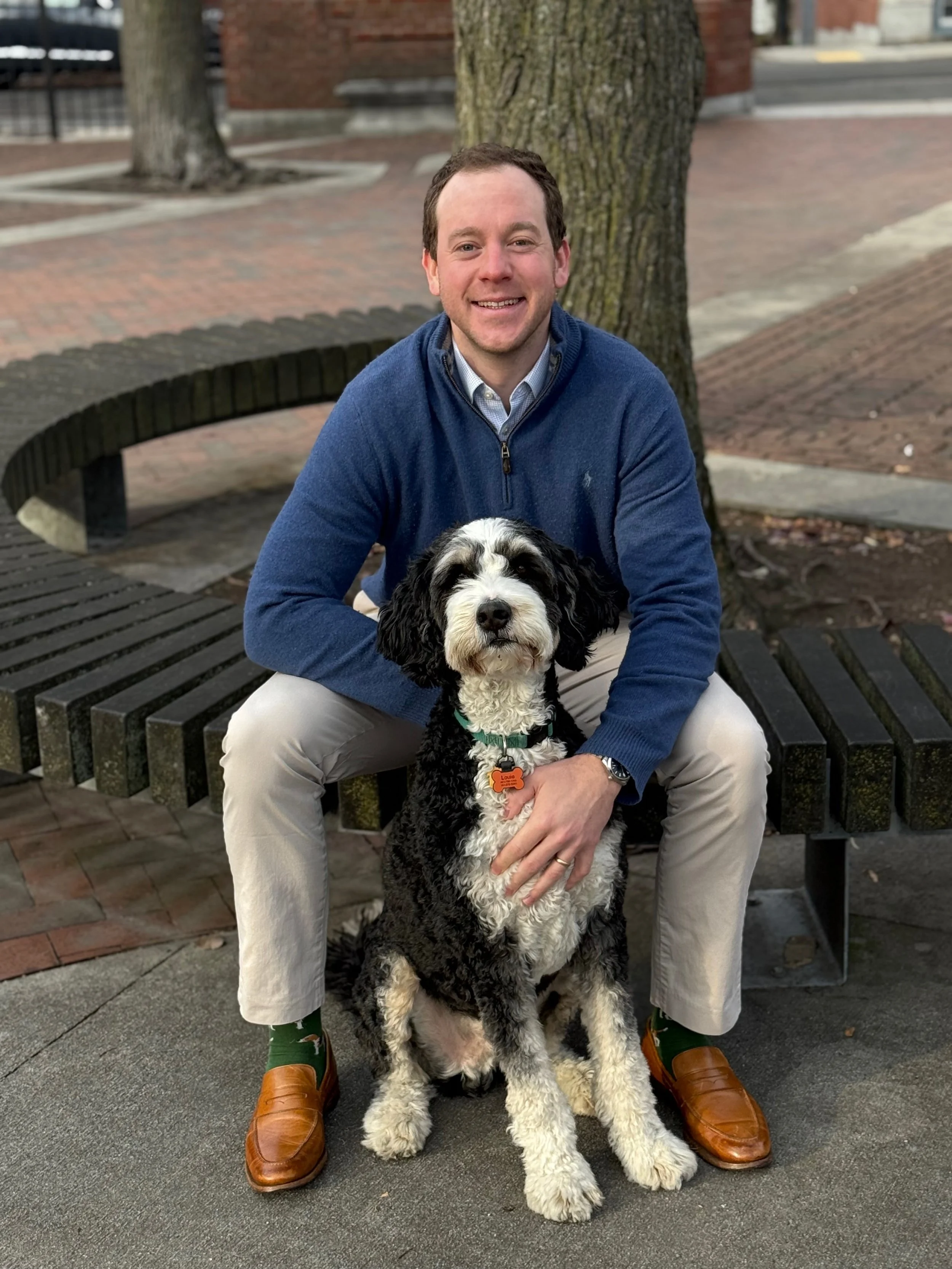 A man in a blue sweater sitting on a park bench with a black and white dog in front of him, outdoors near a tree.