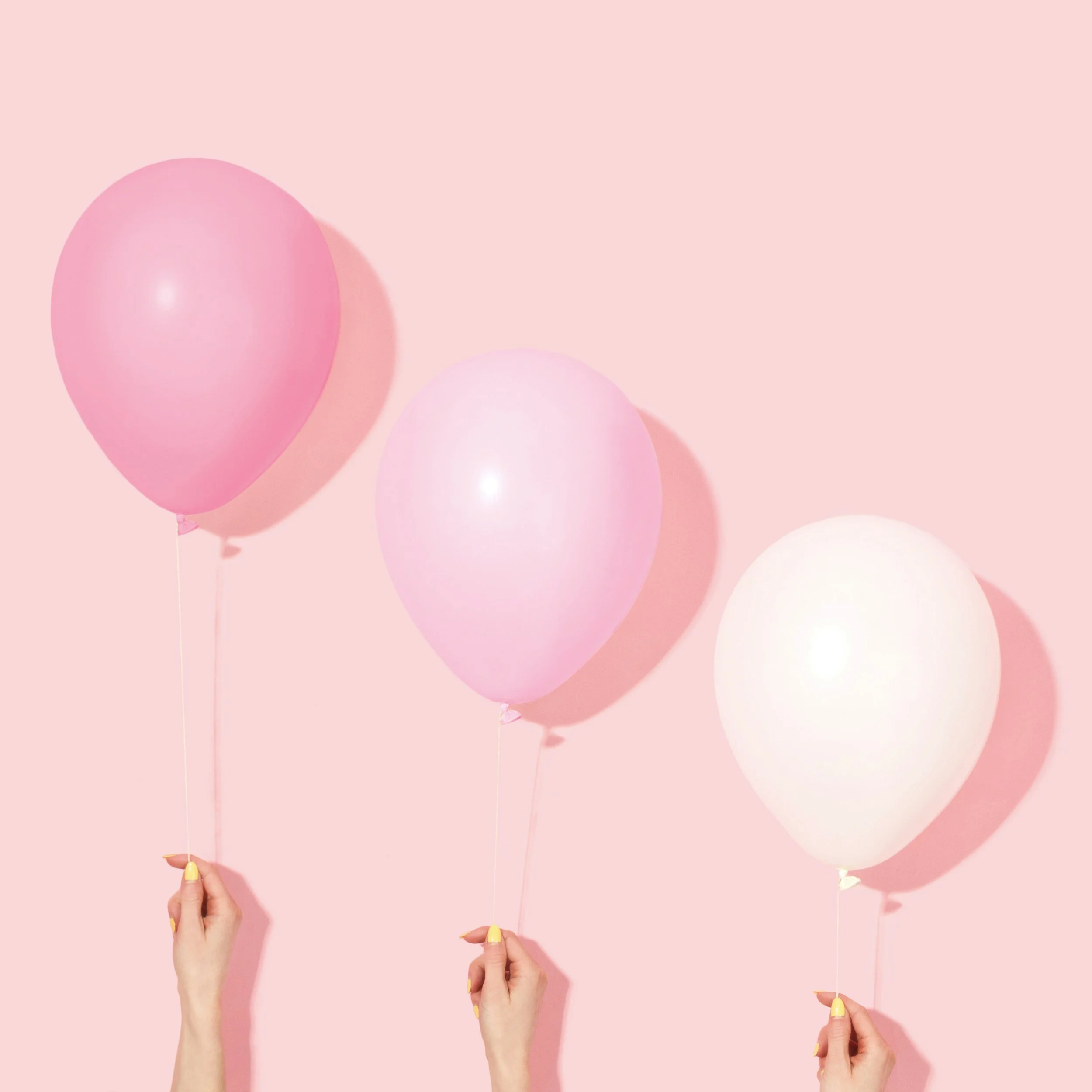 Three hands holding pastel pink, light pink, and white balloons against a pink background.
