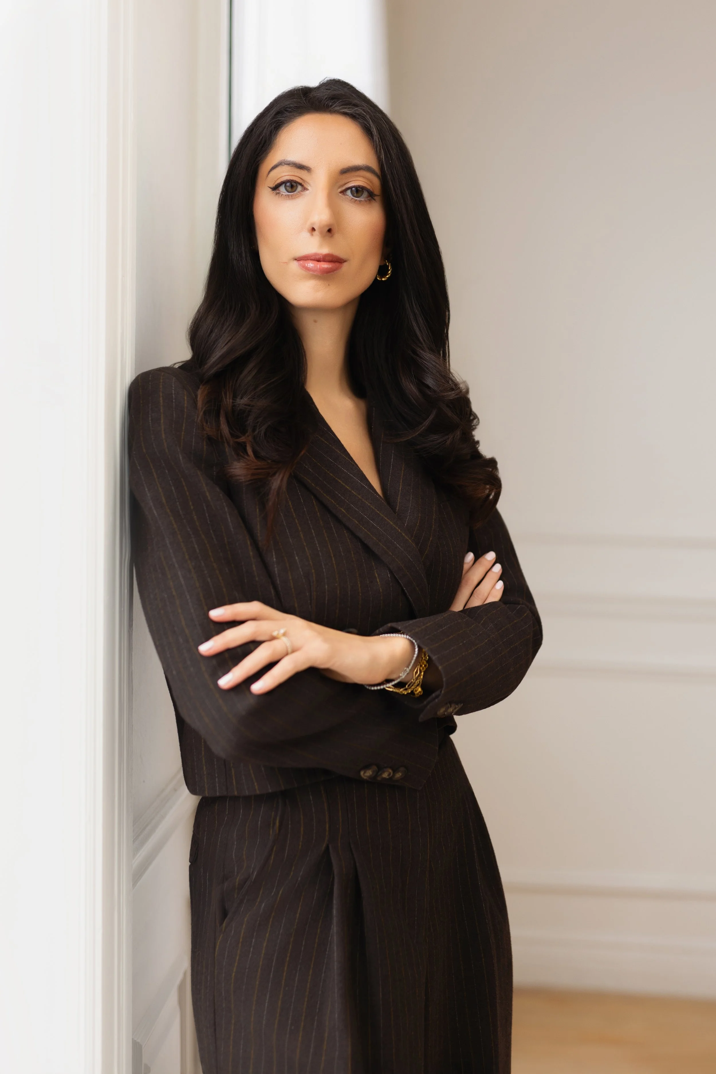A woman with long dark hair, wearing a dark pinstripe suit, standing with arms crossed against a light-colored wall.