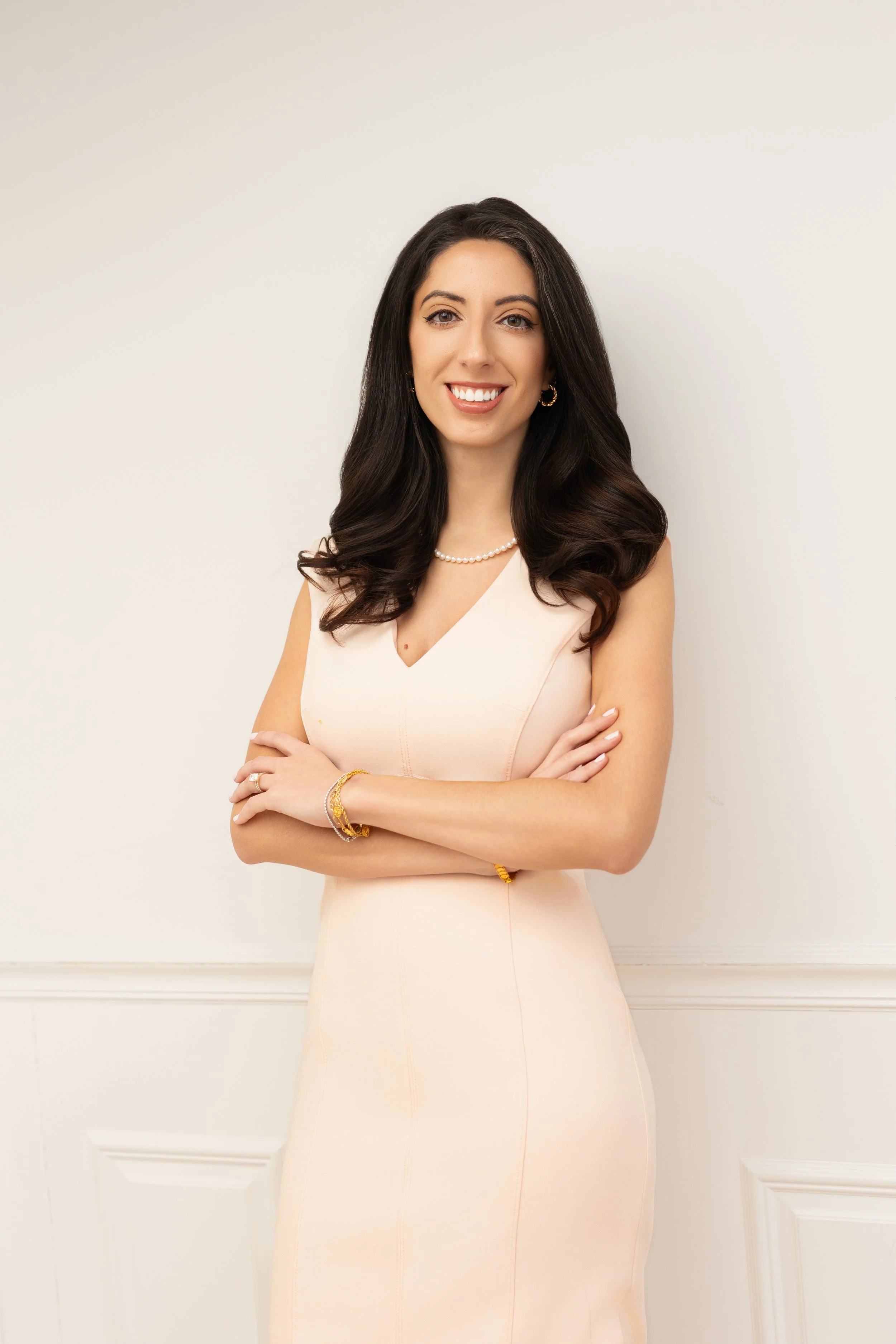 A woman with long dark hair smiling and standing with arms crossed in a pale pink dress against a white background.