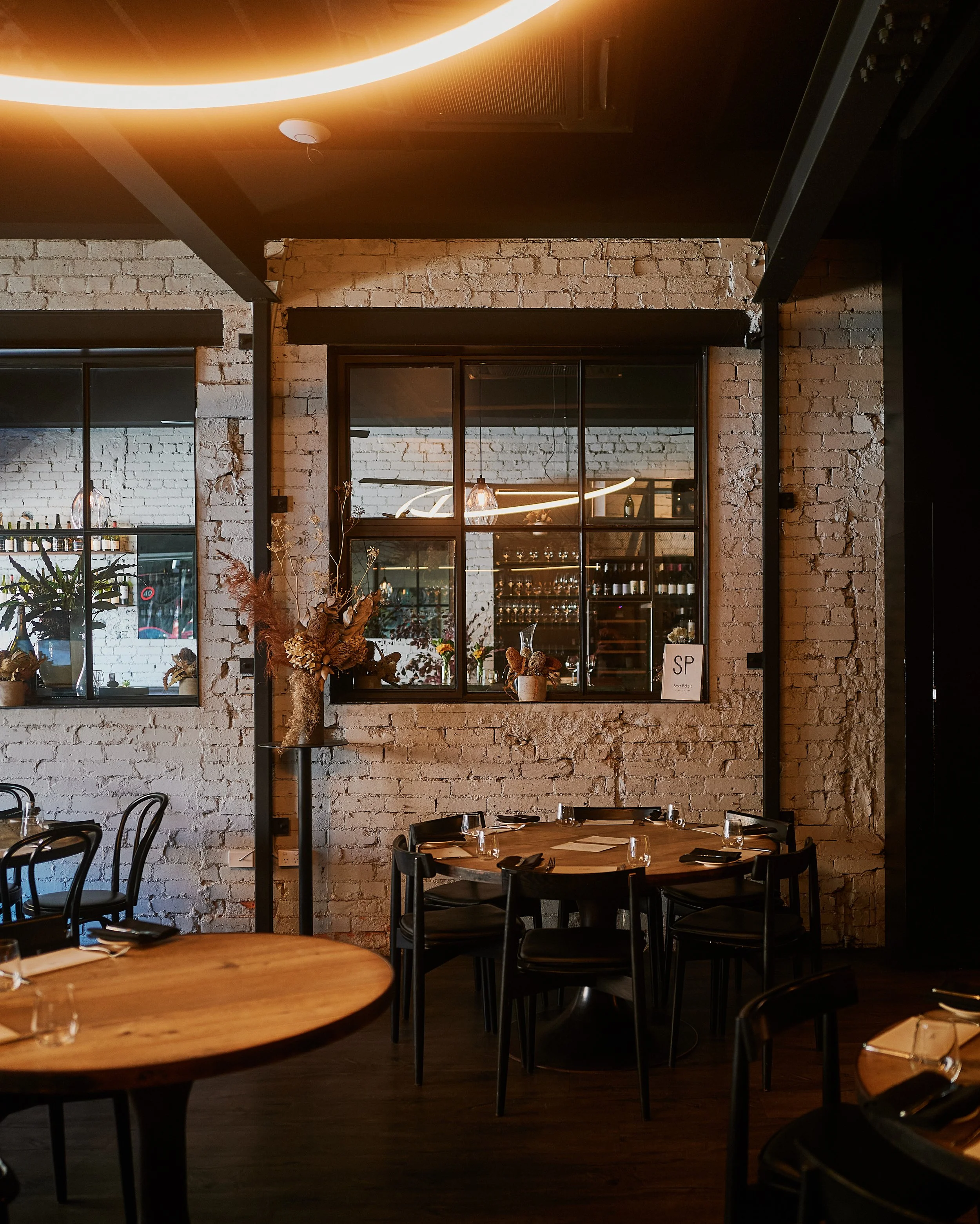 Interior of Estelle with white brick walls, black chairs, wooden tables set for dining, and decorative flower arrangements on a stand and ledge. Large windows and warm lighting create a cozy atmosphere.