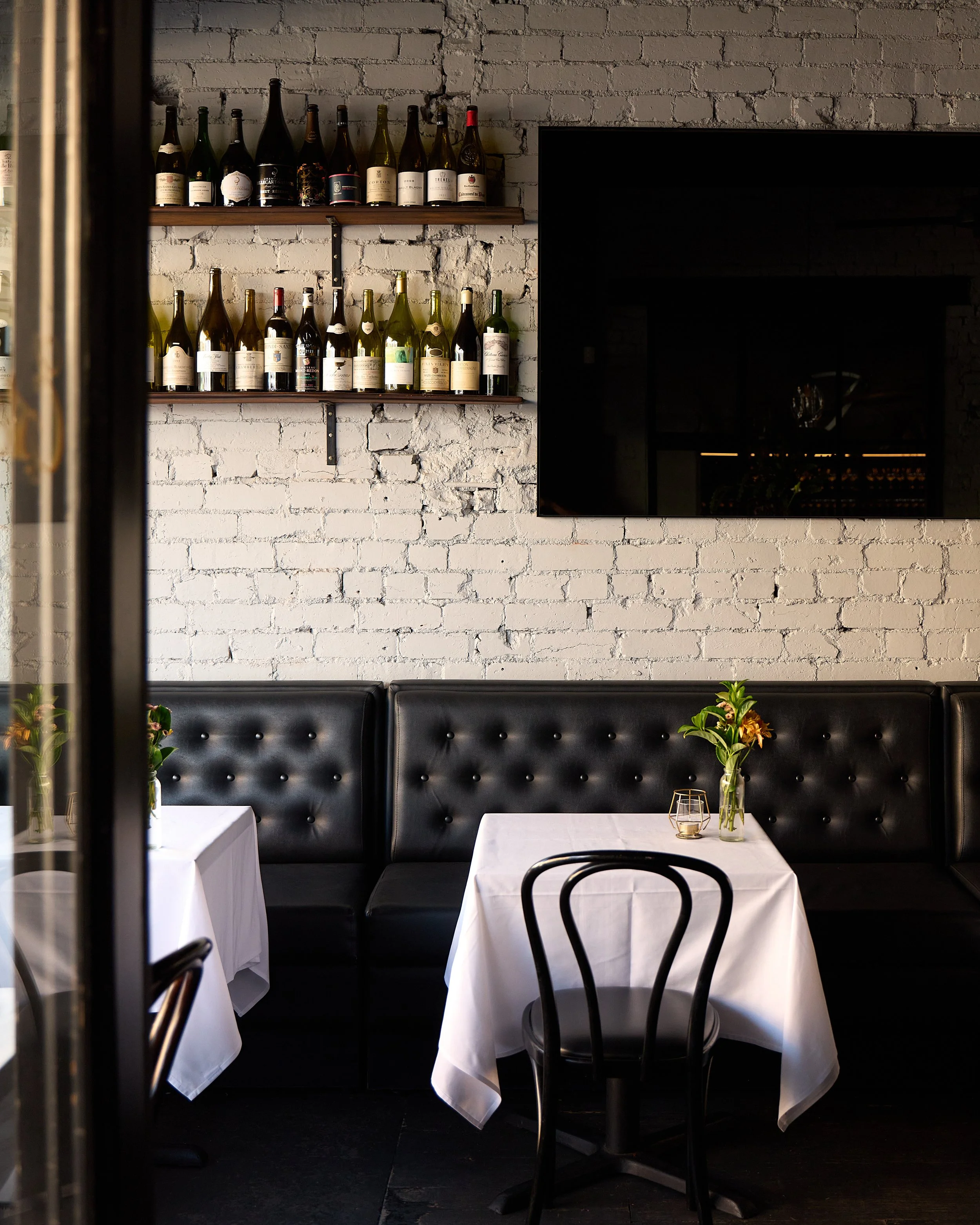 Estelle's interior with white brick walls, black leather banquette seating, and white tablecloth-covered tables decorated with small vases of flowers.