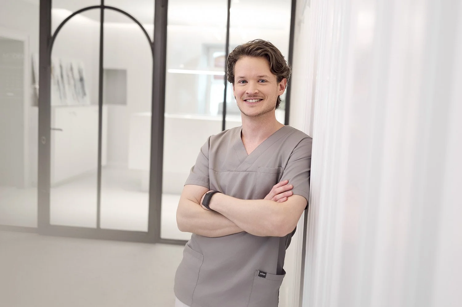 Ein junger Mann in grauen medizinischen Klamotten steht lächelnd in einem hellen, modernen Klinikflur mit Glaswänden.