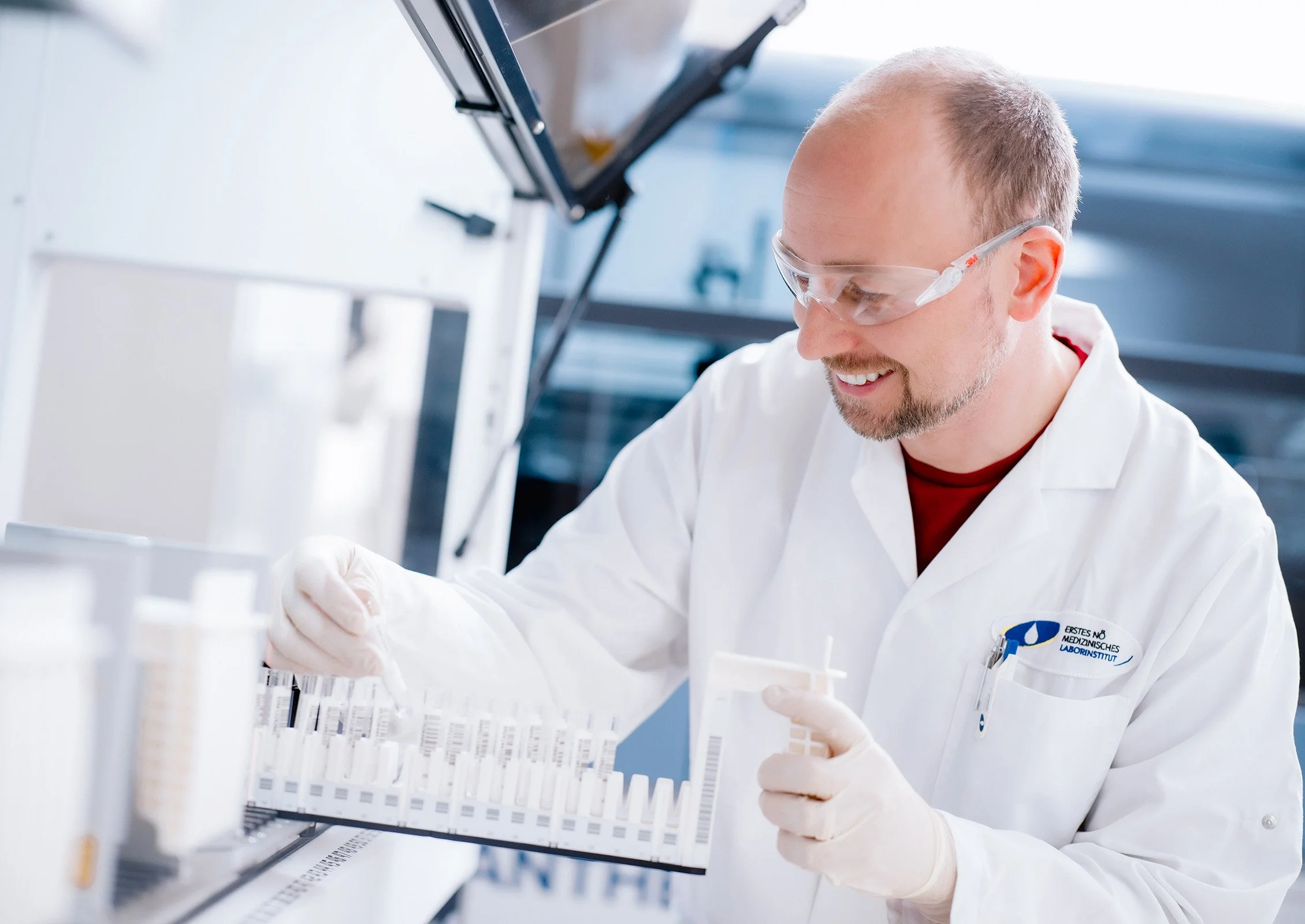 Ein Mann in einem weißen Laborkittel, Schutzbrille und Handschuhen arbeitet in einem Labor mit mehreren Testgefäßen.