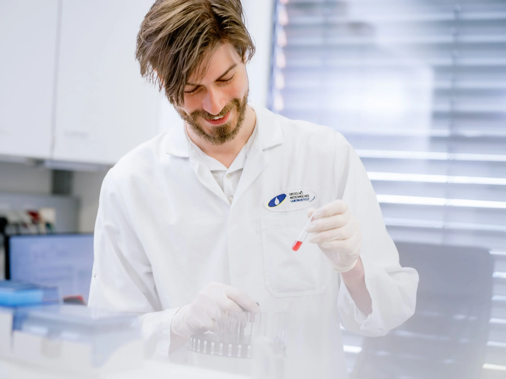 Ein männlicher Wissenschaftler in einem weißen Laborkittel hält eine Probenröhre mit rotem Inhalt und lächelt in einem Labor.