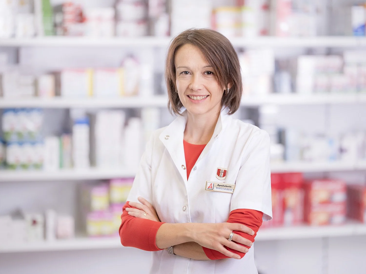Eine Apothekerin steht in einer Apotheke, trägt einen weißen Kittel mit einem Namensschild und hat die Arme verschränkt. Im Hintergrund sind Regale mit Medikamenten und Produkten.