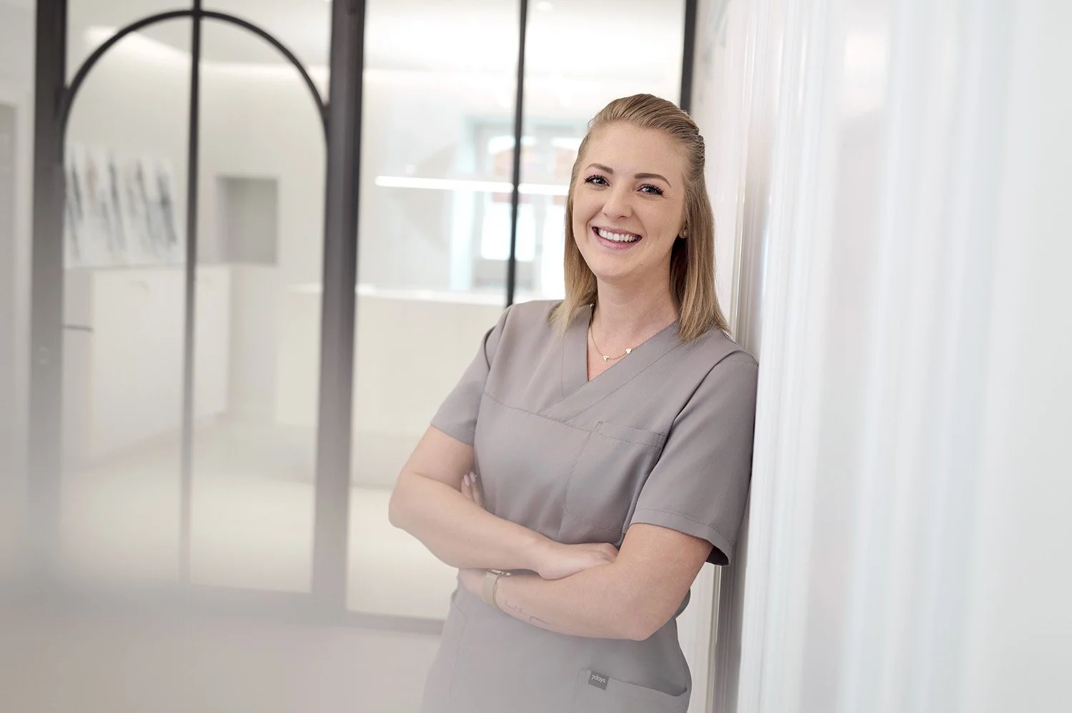 Lächelnde Frau in grauem medizinischen Kittel, die an einer weißen Wand lehnt, im Hintergrund Fenster und Büroeinrichtung.