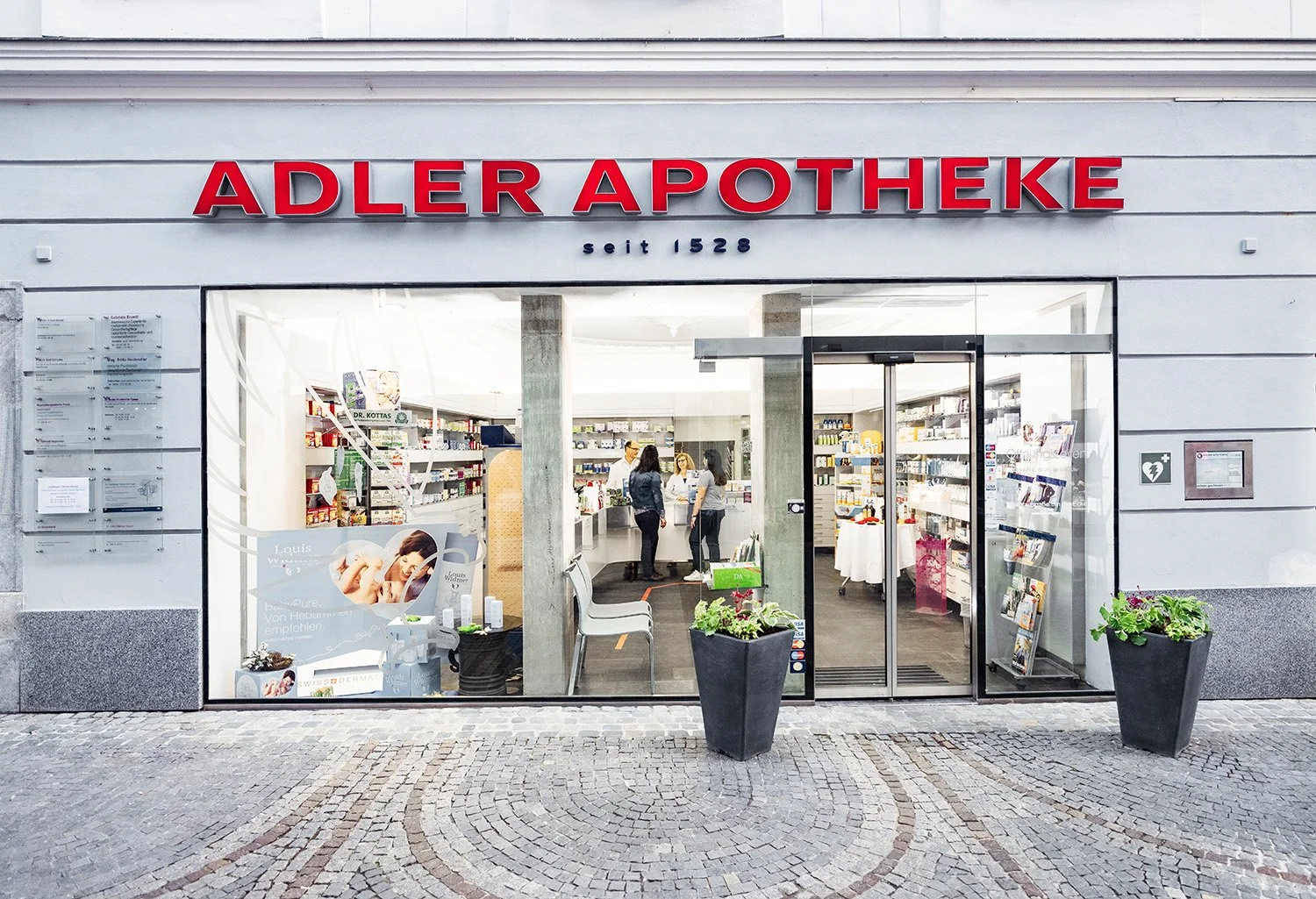 Außenansicht einer Apotheke namens Adler Apotheke mit roten Buchstaben, seit 1528, große Schaufensterfront, zwei große Pflanzgefäße mit Pflanzen vor dem Eingang, Innenansicht mit Kunden und Mitarbeitenden, Regale mit Produkten.