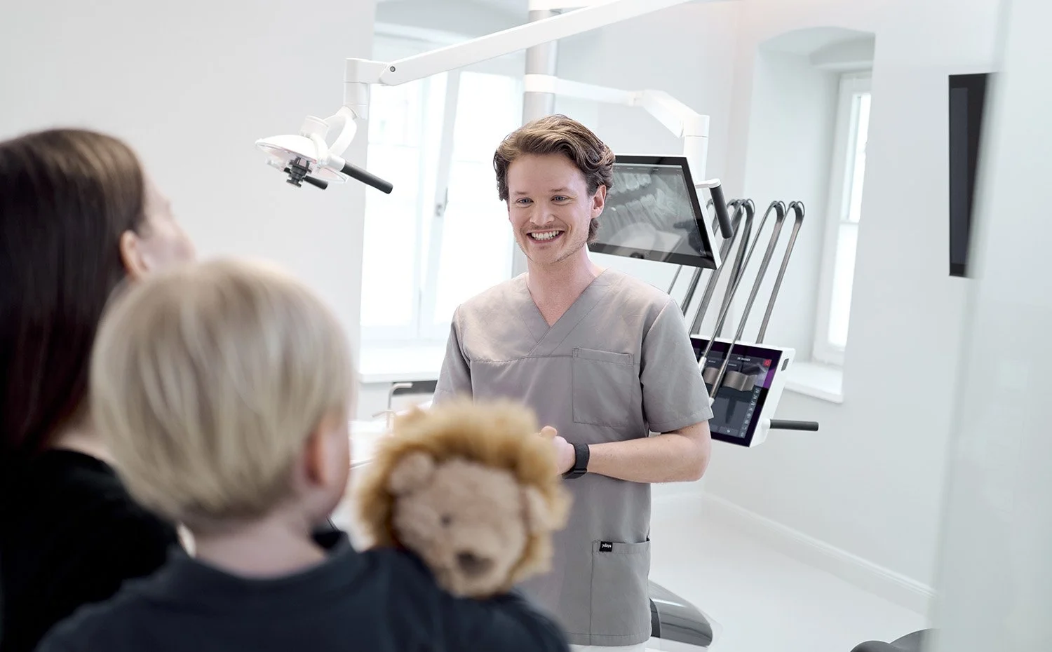 Ein Zahnarzt spricht mit zwei Patienten im Behandlungsraum, im Hintergrund zahnärztliche Geräte und Bildschirme.
