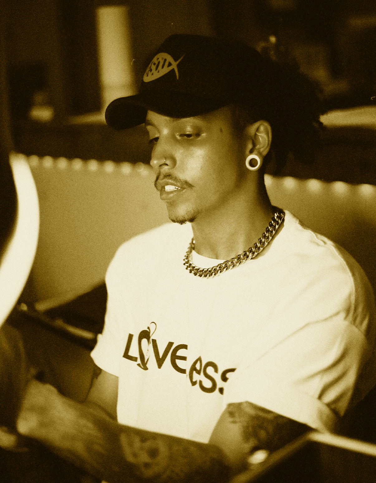 A young man with earrings, a nose ring, and tattoos wearing a cap and a white shirt with the word LOVELESS, sitting at a desk or table in a dimly lit room.