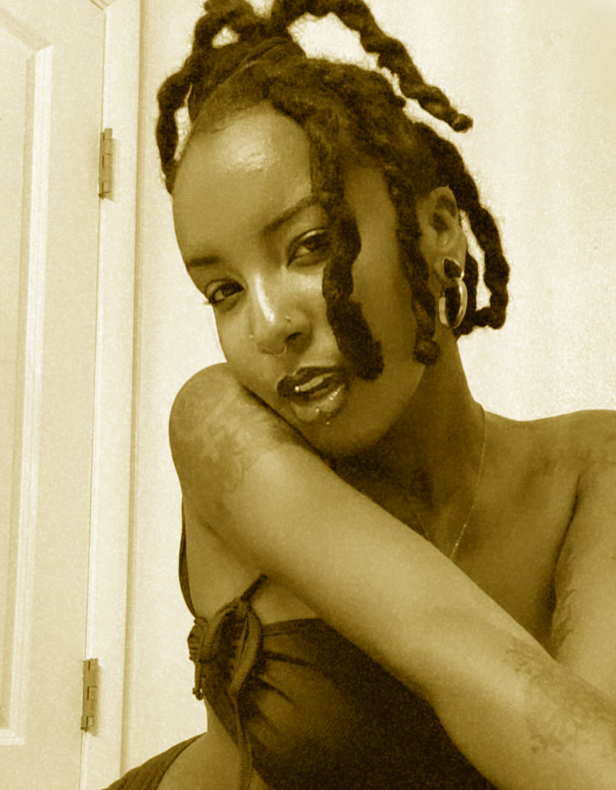 A woman with dreadlocks, multiple earrings, and a lip piercing, leaning her head on her arm and looking at the camera with a relaxed expression.
