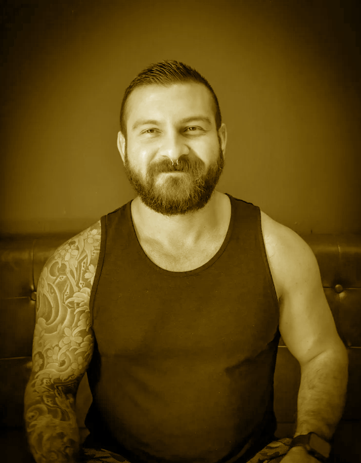 A bearded man with a tattoo on his left arm, wearing a sleeveless shirt, smiling and sitting indoors against a plain background.