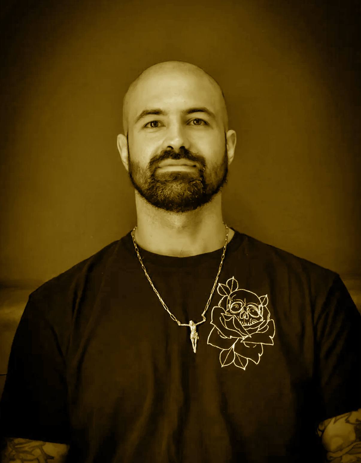 Portrait of a man with a beard, mustache, and shaved head, wearing a chain necklace with a pendant and a T-shirt with a graphic of a skull and rose on it, against a plain background.