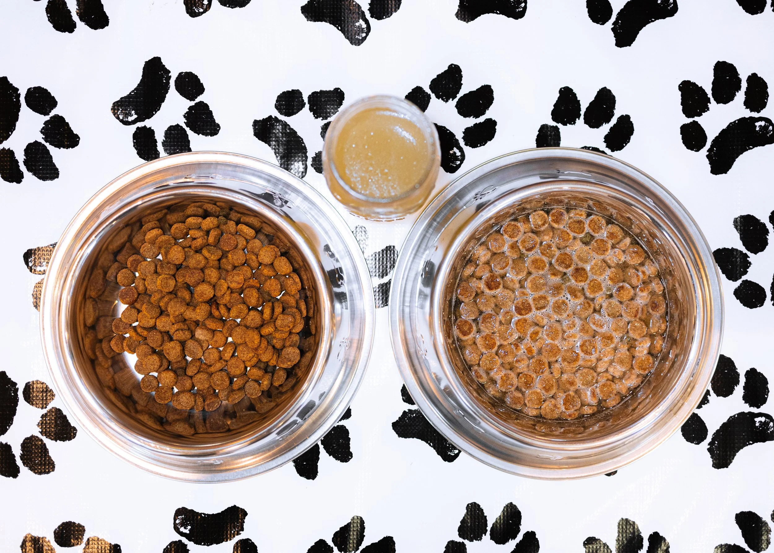 Two glass bowls of dry and soaked dog food with a small glass of water on a table with a black and white paw print pattern.