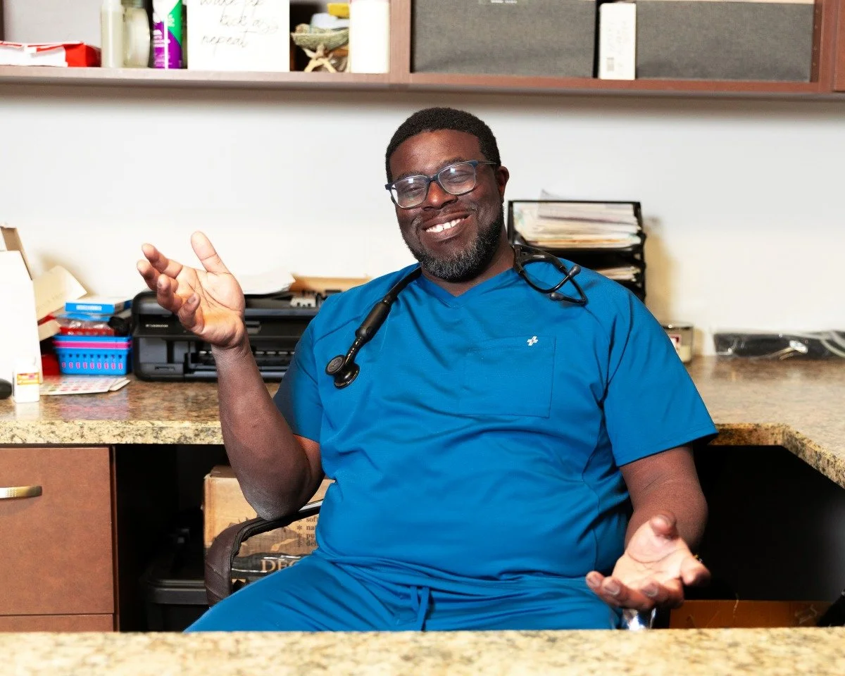Dr Jeremy in blue scrubs with a stethoscope around his neck sitting at a cluttered desk or counter in an office or clinic room.