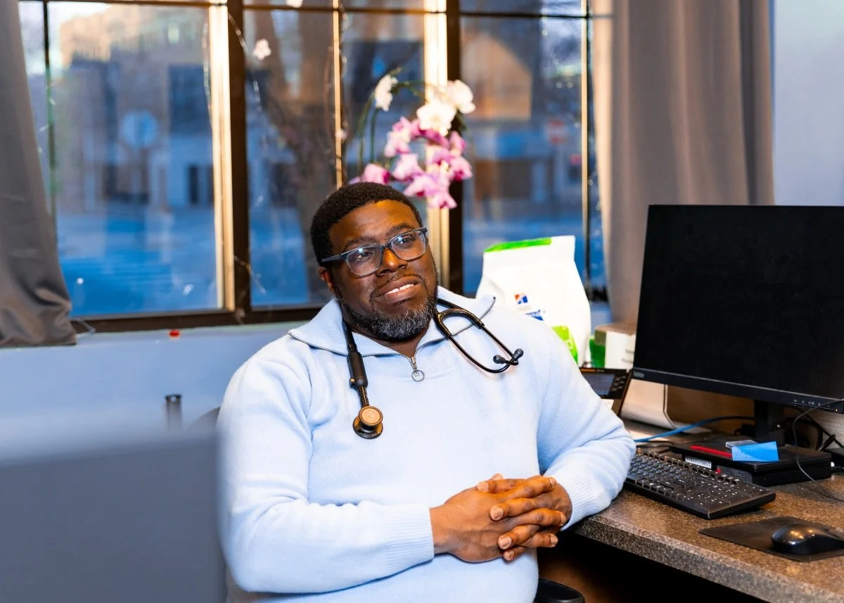 Dr Jeremy wearing a light blue sweater and a stethoscope, is sitting at a desk in an office. There are medical supplies in the background, and a large computer monitor to his right. The window behind him shows some trees and buildings outside.