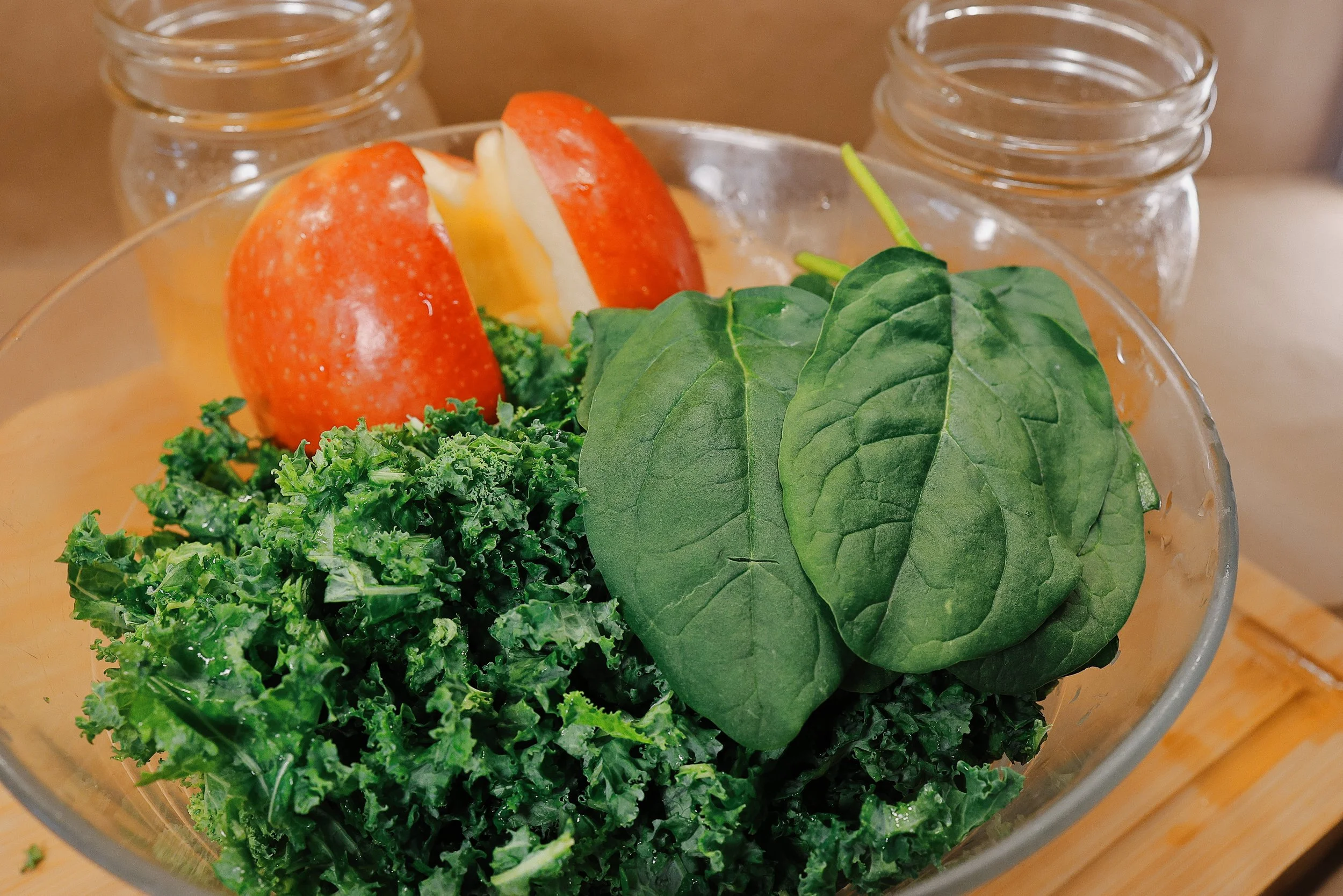 A glass bowl filled with leafy green vegetables, including spinach and kale, and a red apple with a bite taken out of it, on a wooden surface.