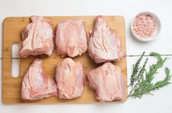 Six raw pork shoulder cuts on a wooden cutting board with herbs, pink Himalayan salt, and a bowl of pink Himalayan salt nearby.