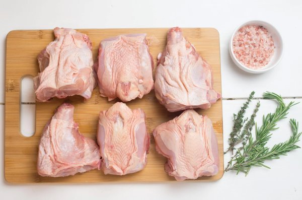Six pieces of raw pork on a wooden cutting board with a bowl of pink Himalayan salt and sprigs of fresh herbs including rosemary and thyme on a white surface.