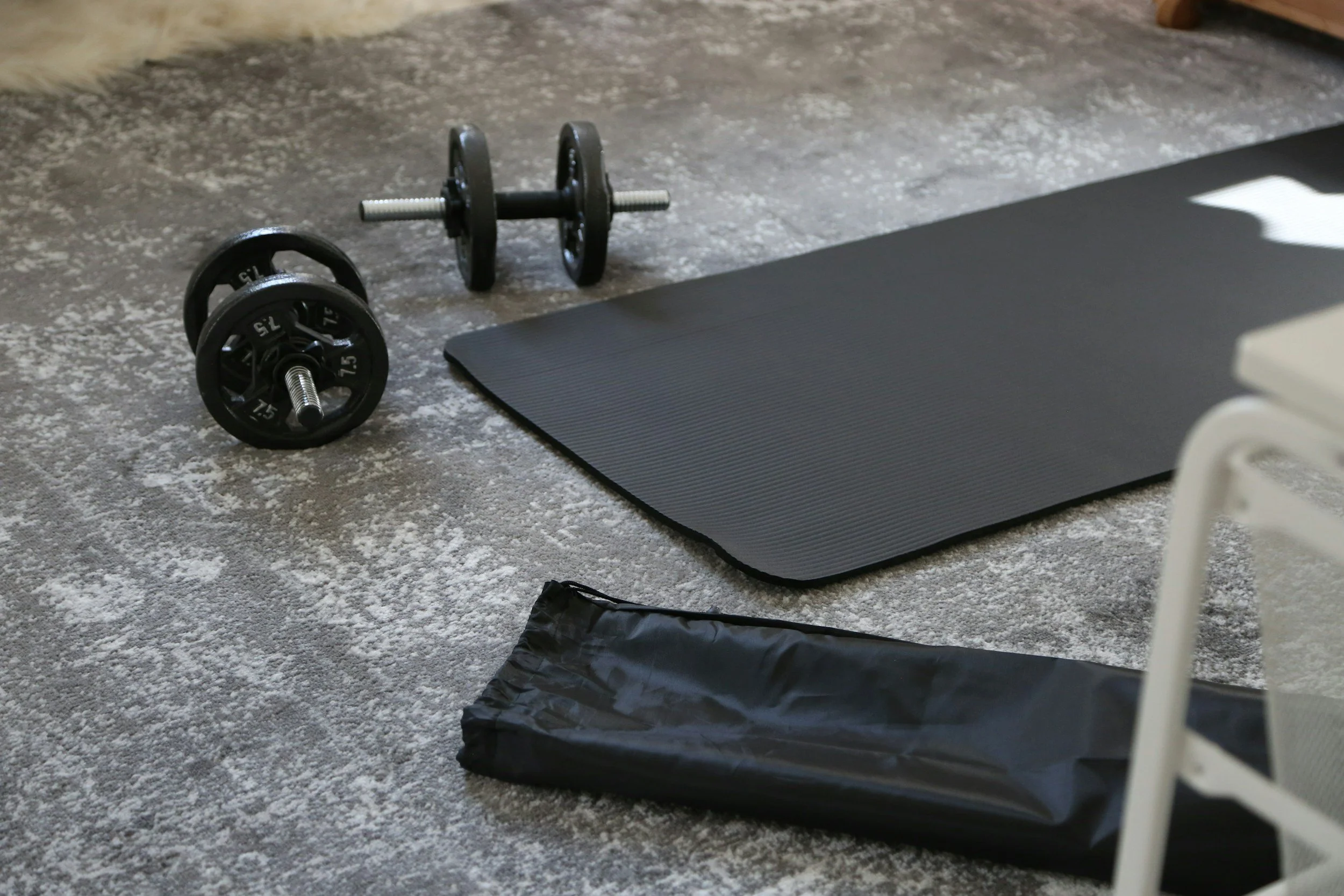 Dumbbells, a yoga mat, a folded exercise mat, and a chair are on a padded floor, indicating a home gym setup.
