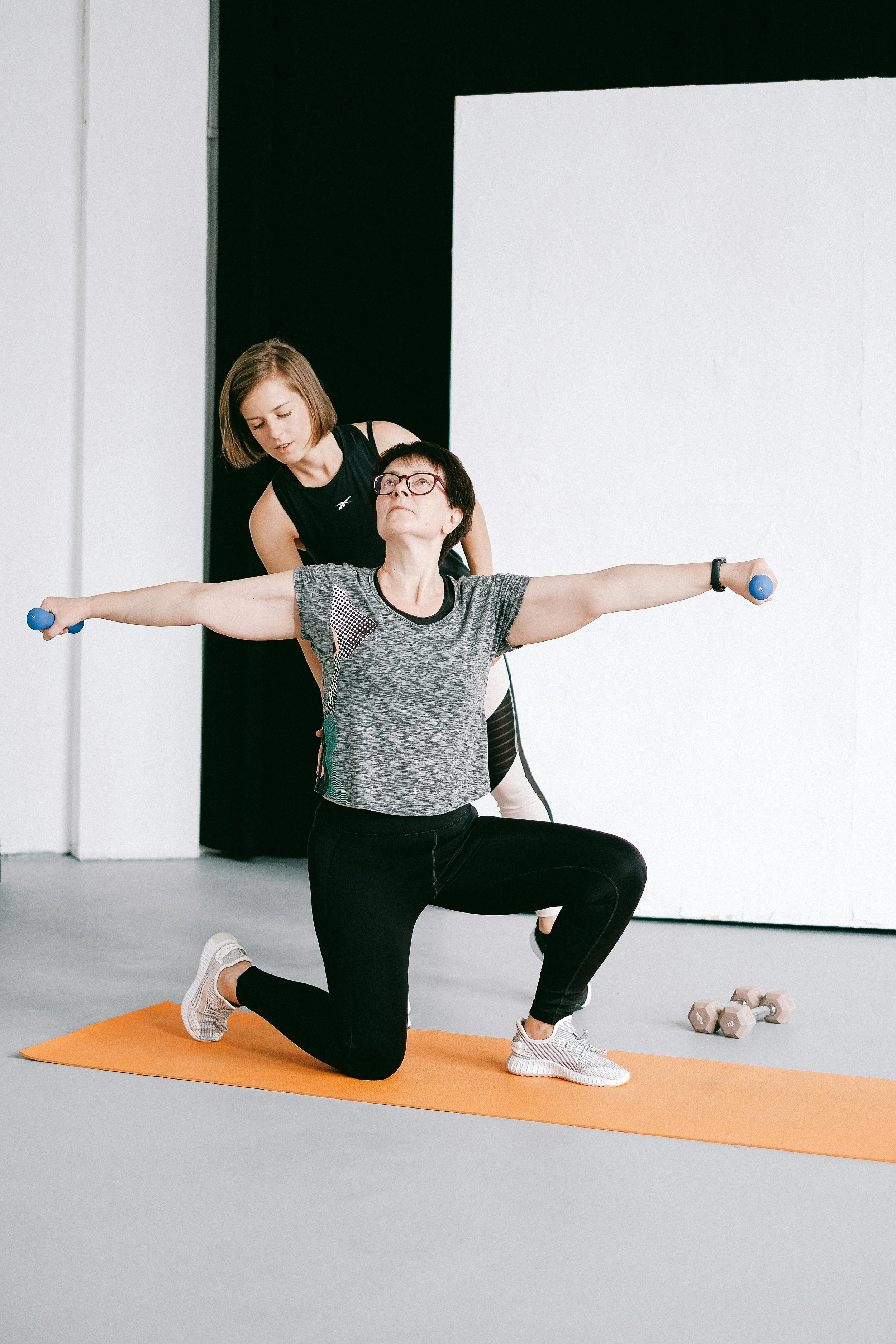 To kvinder træner i et fitnesscenter. Den ene laver en knæbøjning med håndvægte, mens den anden overvåger og guider hende.