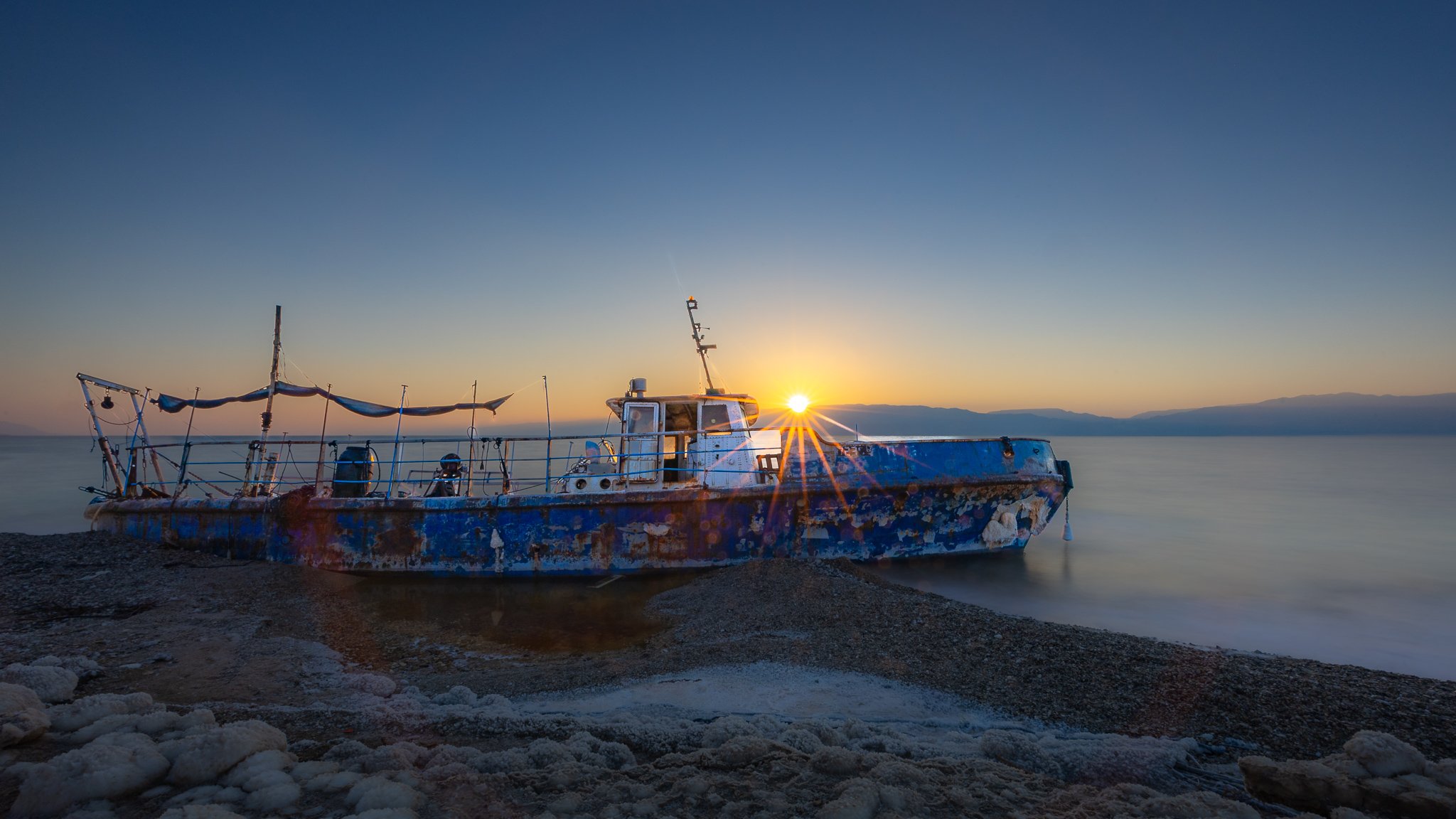 Abandoned blue boat on rocky shore at sunset with calm water and distant mountains.
