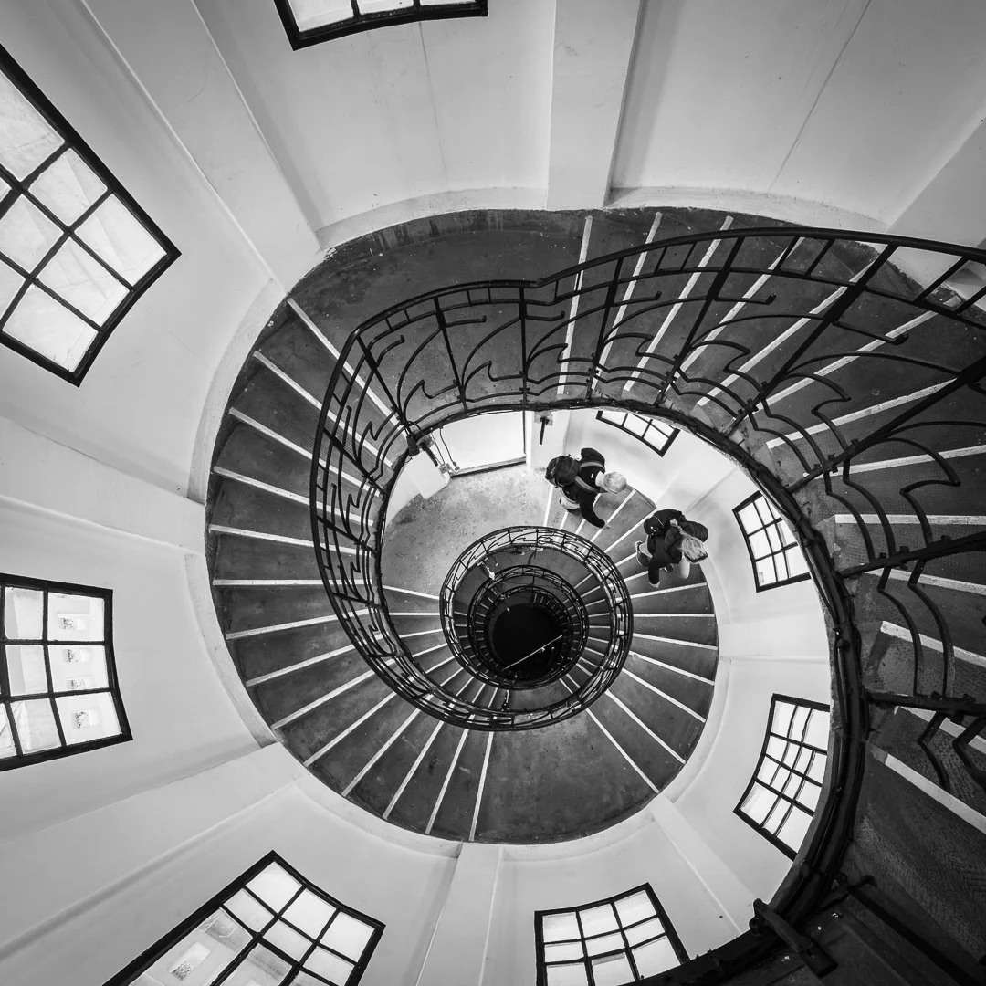 Spiral staircase viewed from above, with two people walking down, black railing, white walls, and windows.