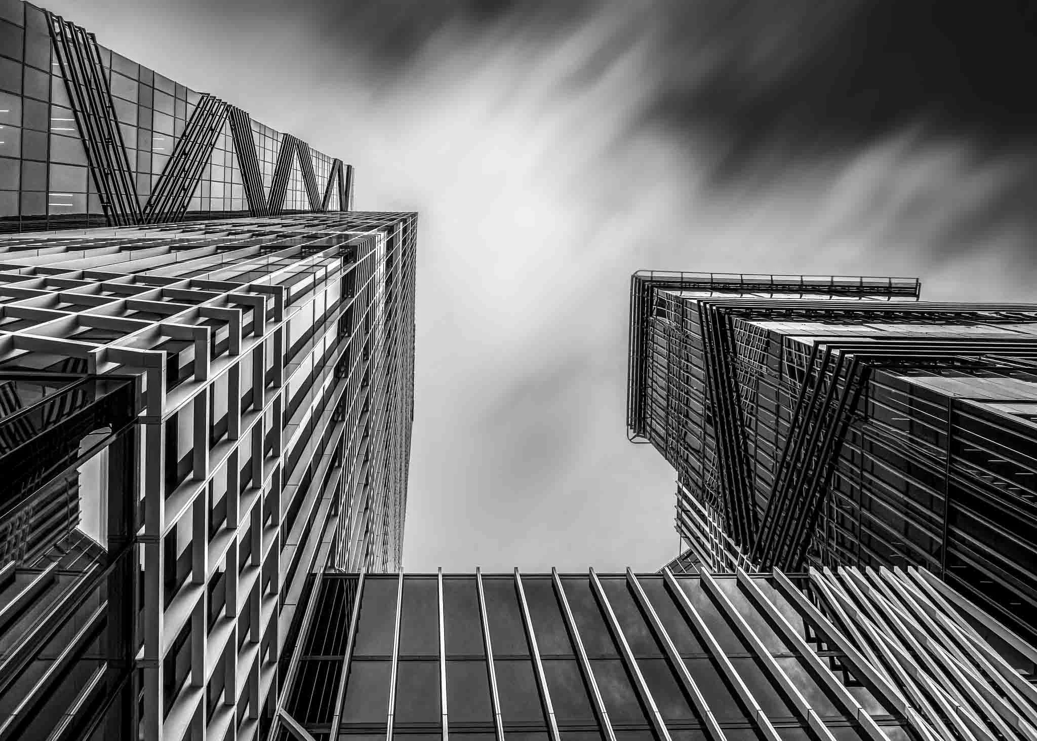Black-and-white fine-art photograph looking upward between modern high-rise buildings, with dramatic blurred clouds emphasizing the geometric architecture.