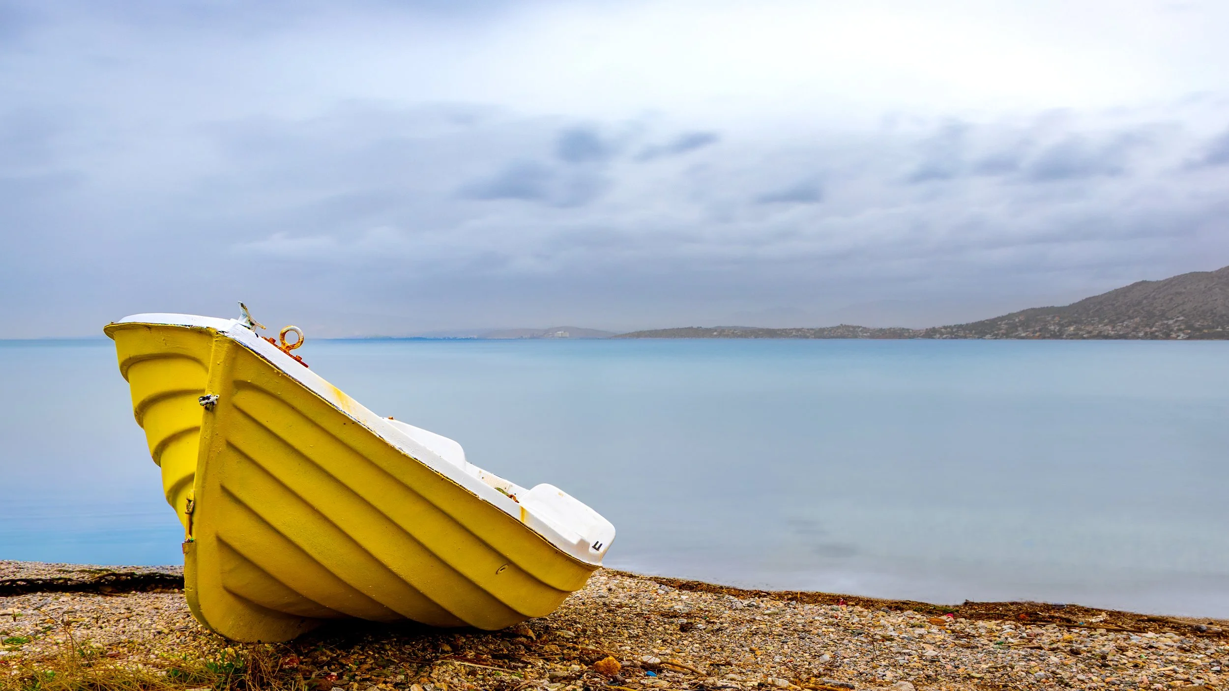 Yellow boat resting on a rocky beach with calm water and cloudy sky in the background, photograph by erez nudmanov