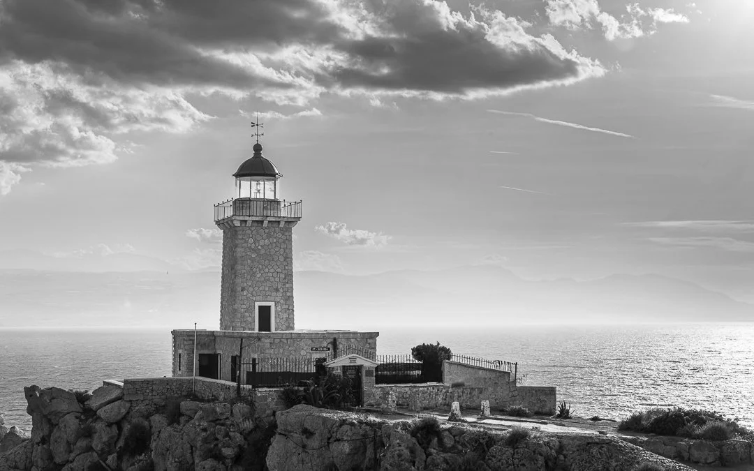 A stone lighthouse on a rocky shoreline overlooking the ocean with clouds in the sky.