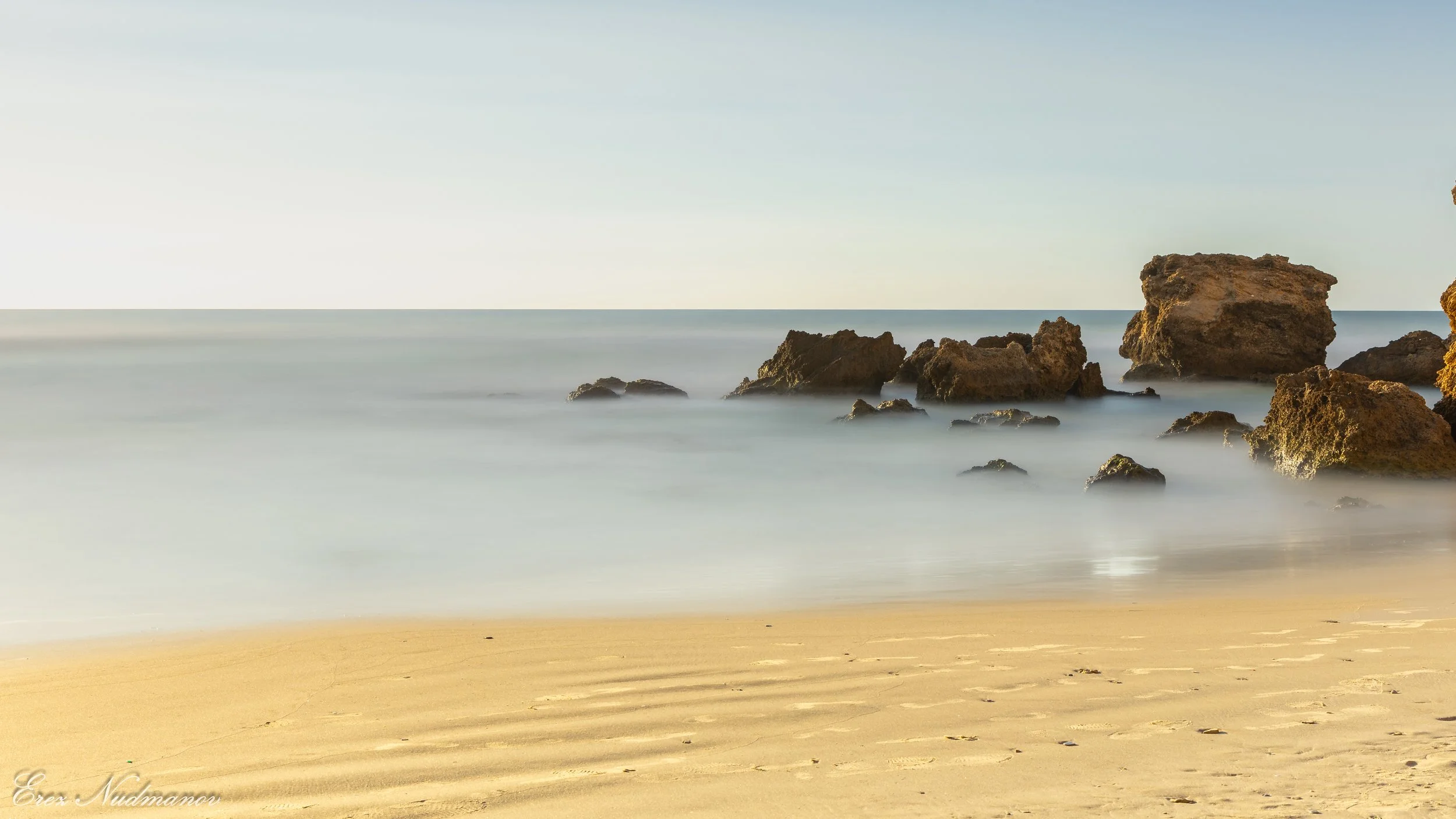 A beach scene with sandy shore, rocks in the ocean, and a calm sky.