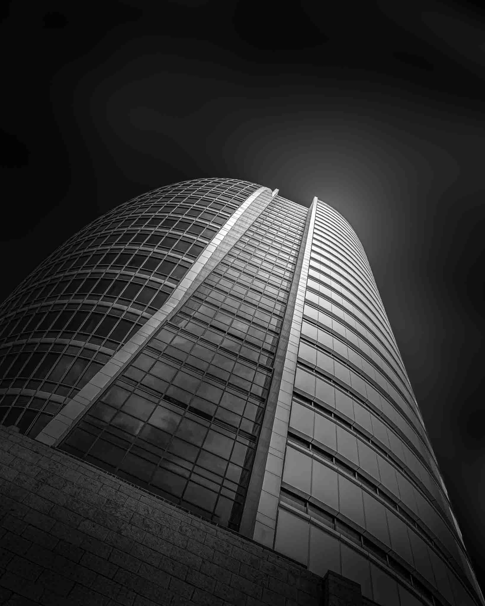 Black and white fine art photograph of Haifa’s Sail Tower from a dramatic low-angle perspective, emphasizing its curved glass facade and geometric lines.