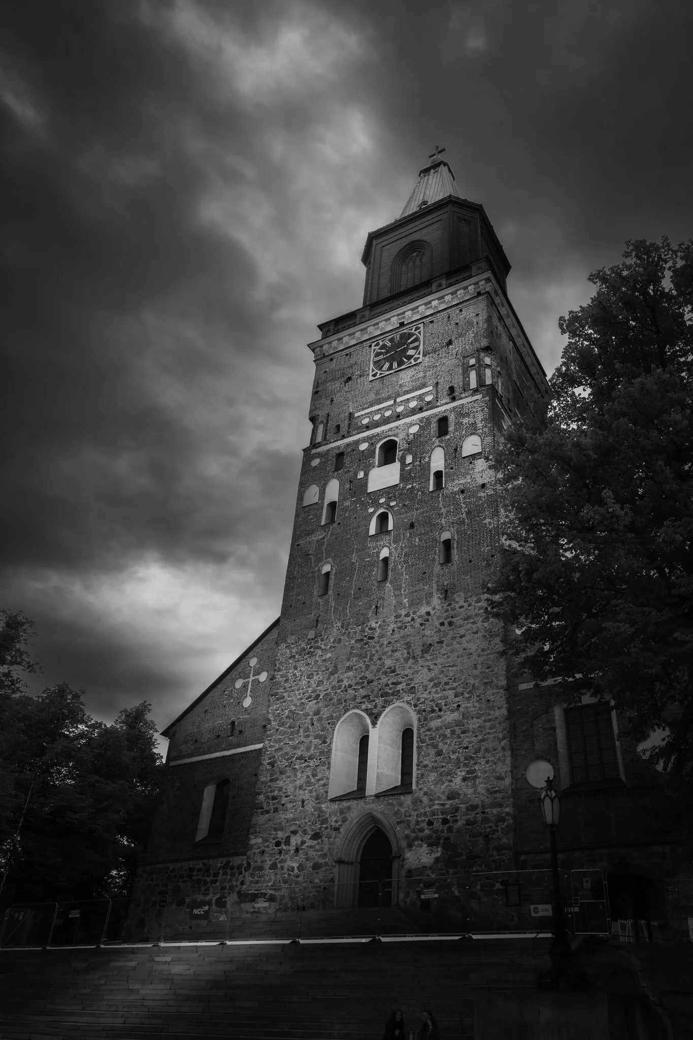 A tall stone church with a clock on its front facade, a steeple with a cross at the top, and Gothic-style windows, set against a cloudy sky, with trees around the building.