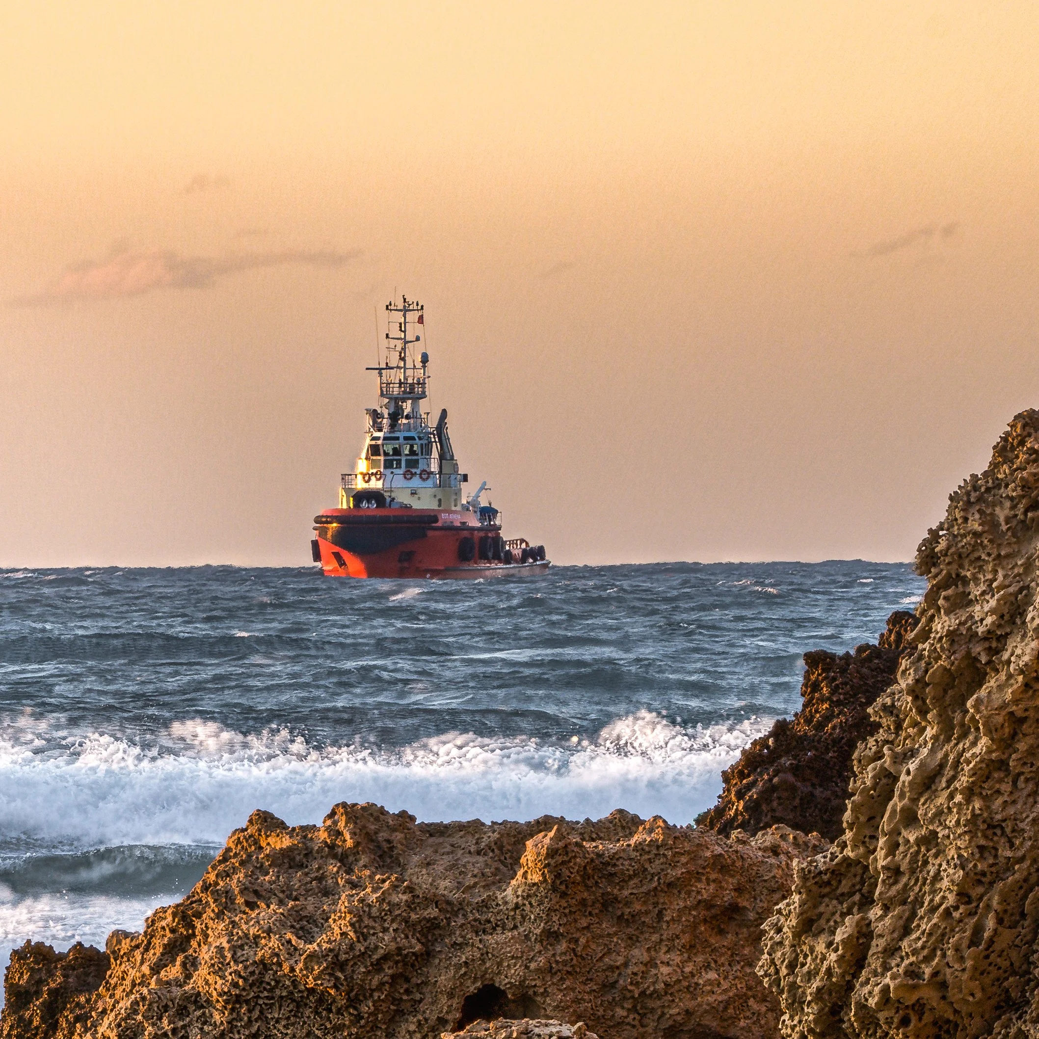 A boat sailing on the ocean with rocky shoreline in the foreground at sunset.