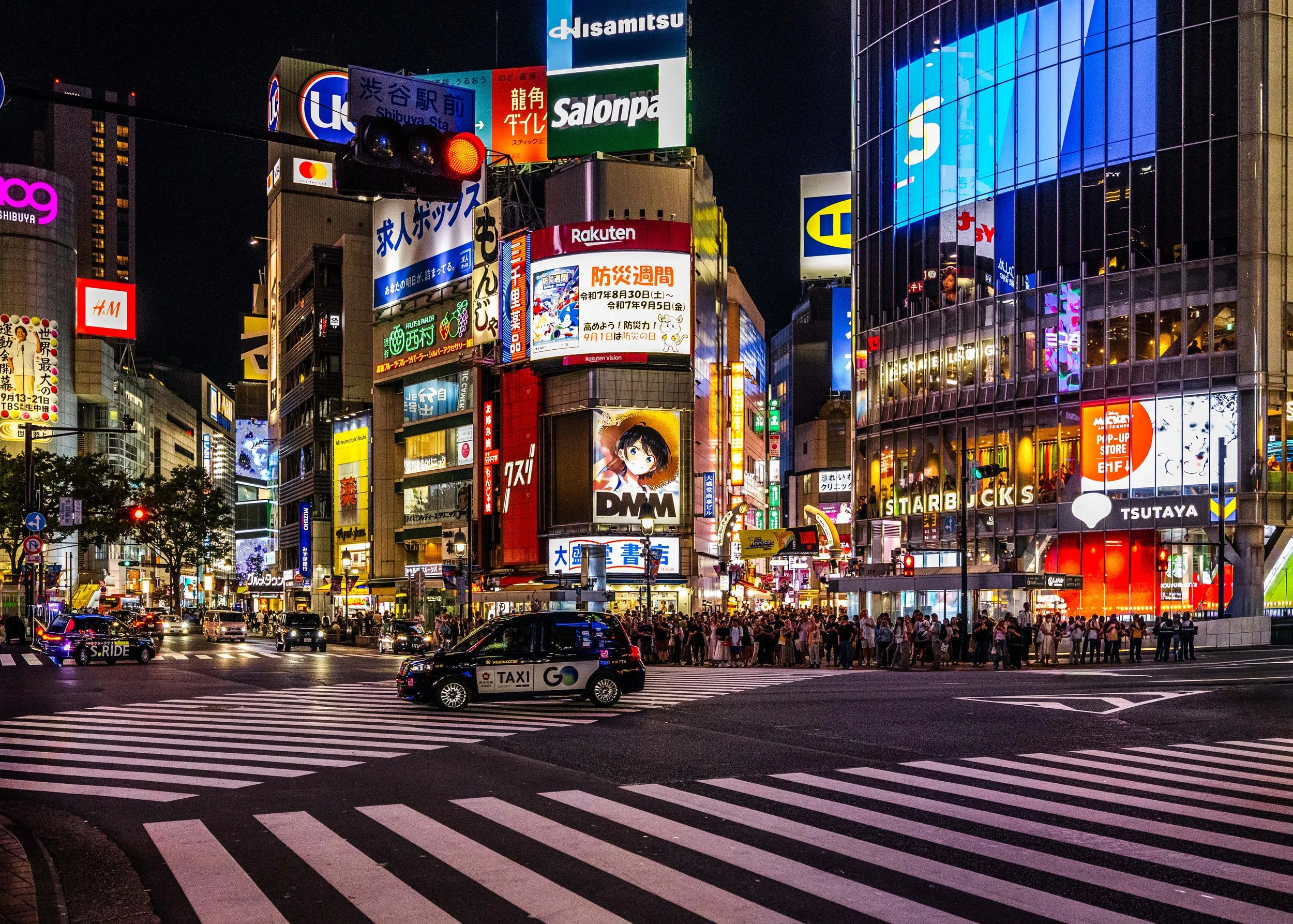 Nighttime city scene with illuminated billboards, neon signs, and a crowd at a busy intersection in Tokyo, Japan.