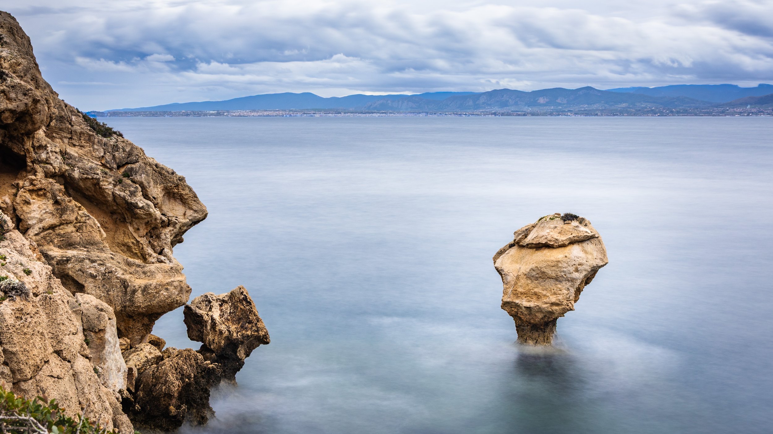 Rock formation along a calm sea, with a large rock protruding from the water and cliffs on the left, distant mountains in the background, and a cloudy sky, photograph by erez nudmanov