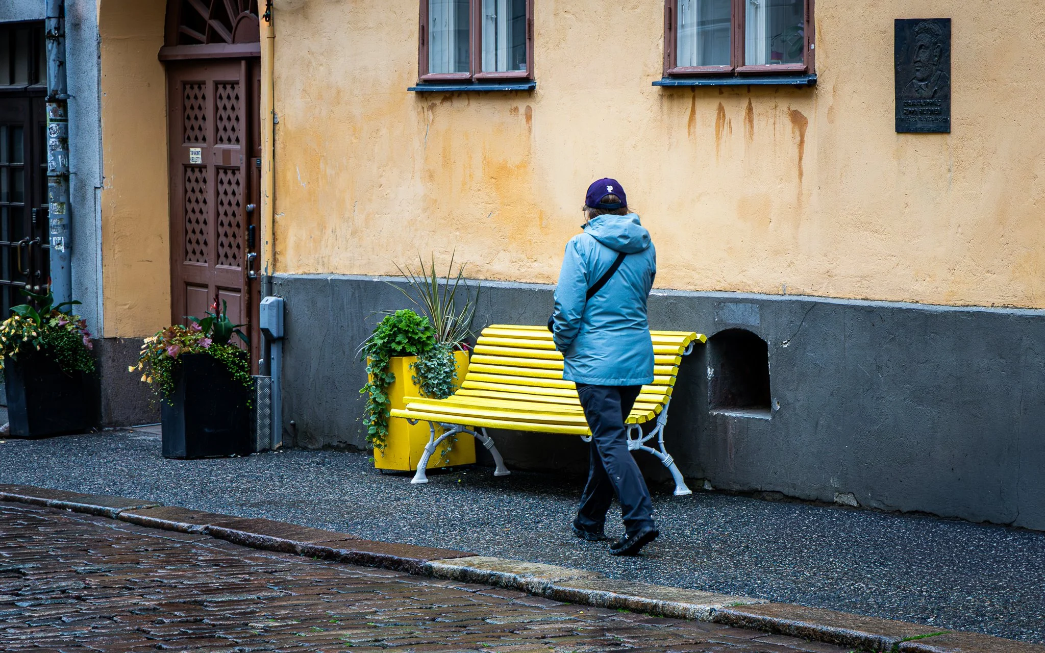 A person in a blue jacket and black pants walking past a yellow park bench on a city sidewalk. The person is wearing a navy cap and glasses, facing away from the camera. There are two windows on a yellow building and potted plants in black containers