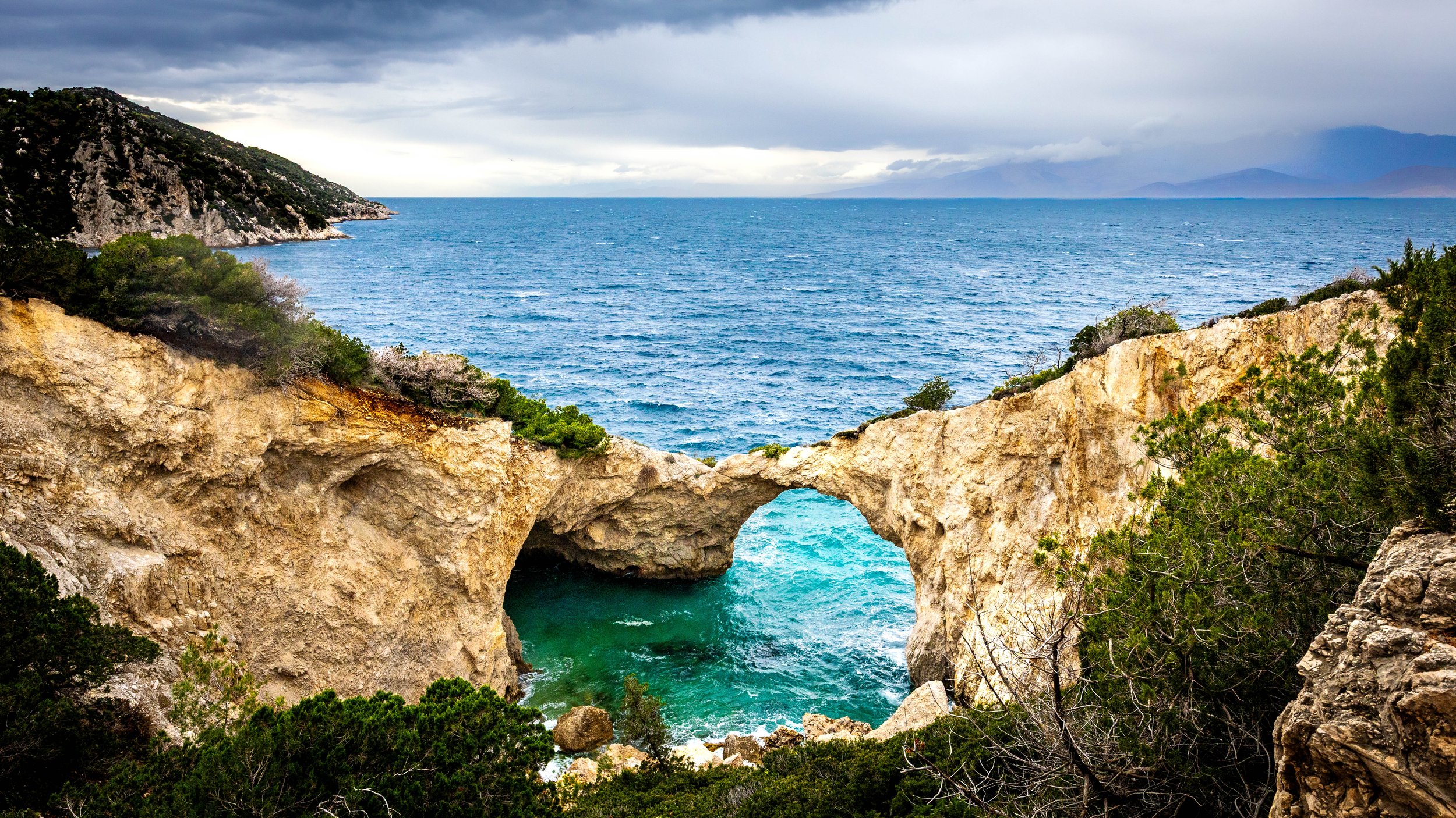 Rock formation with a natural arch over turquoise water on a rugged coastline, with the ocean extending into the distance under cloudy skies.
